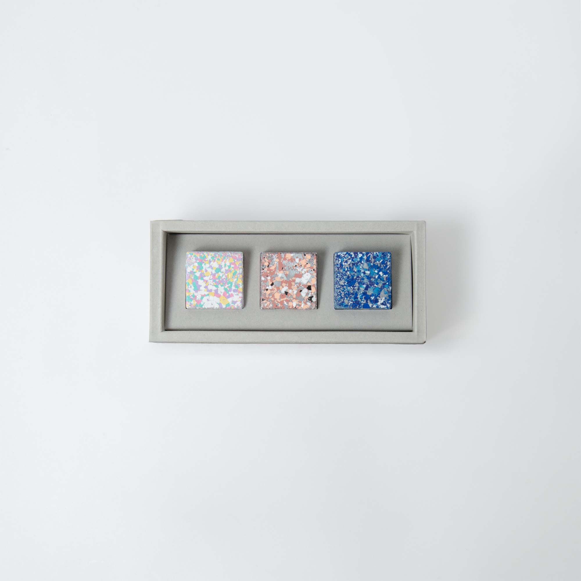 Das Set „Color Gem – das Wachsmalquadrat“ von Studio Unto enthält drei Glitzerpfannen in schillerndem Weiß, Roségold und Blau, angeordnet in einer rechteckigen Box auf einem schlichten weißen Hintergrund.