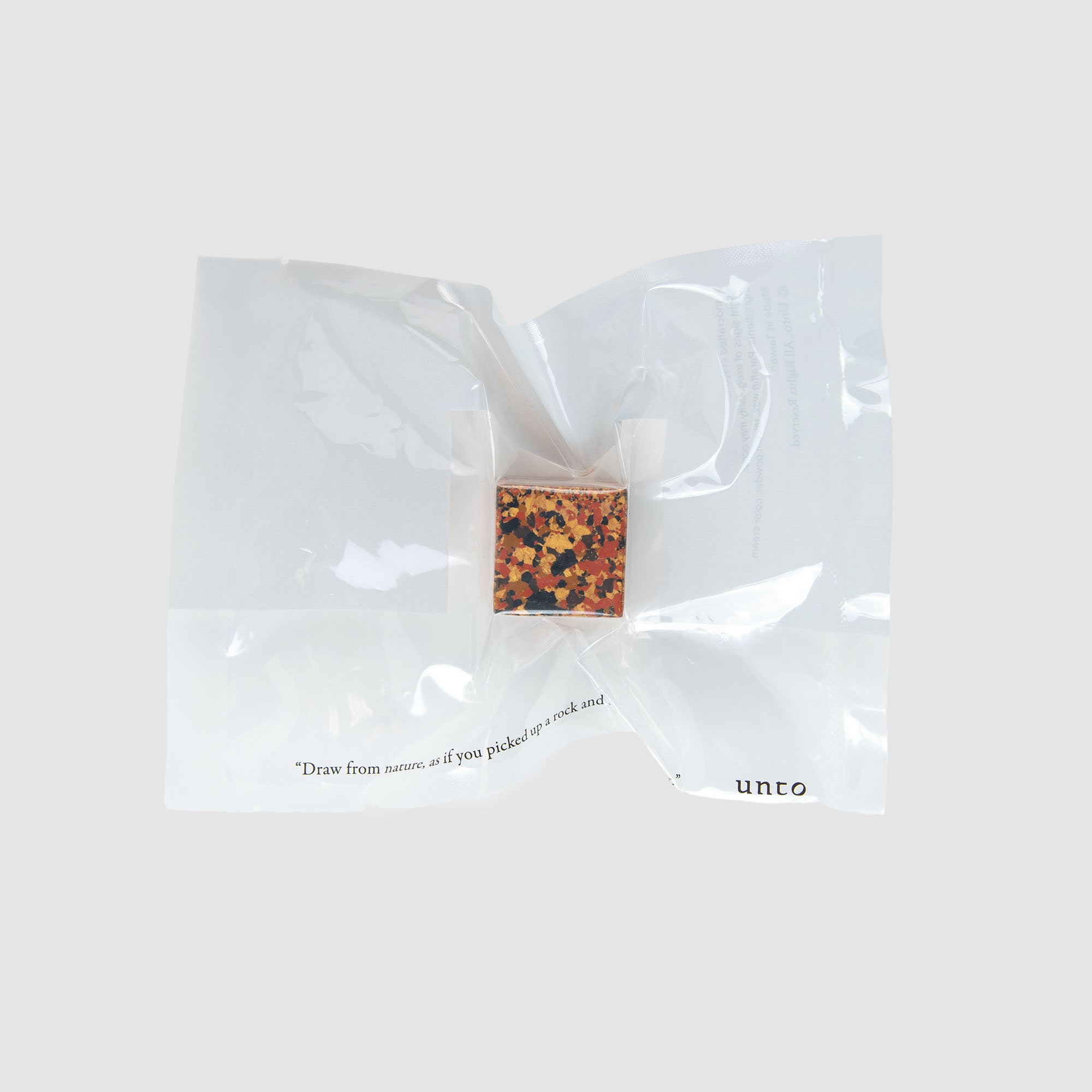 Eine kleine, farbenfrohe, abstrakte Schachtel namens „Color Gem – das Wachsmalquadrat – Einzelpackung“ von Studio Unto liegt auf einer durchscheinenden, zerknitterten Plastikfolie. Ein Teil des Textes auf der Folie enthält das Wort „unto“ vor einem schlichten weißen Hintergrund.