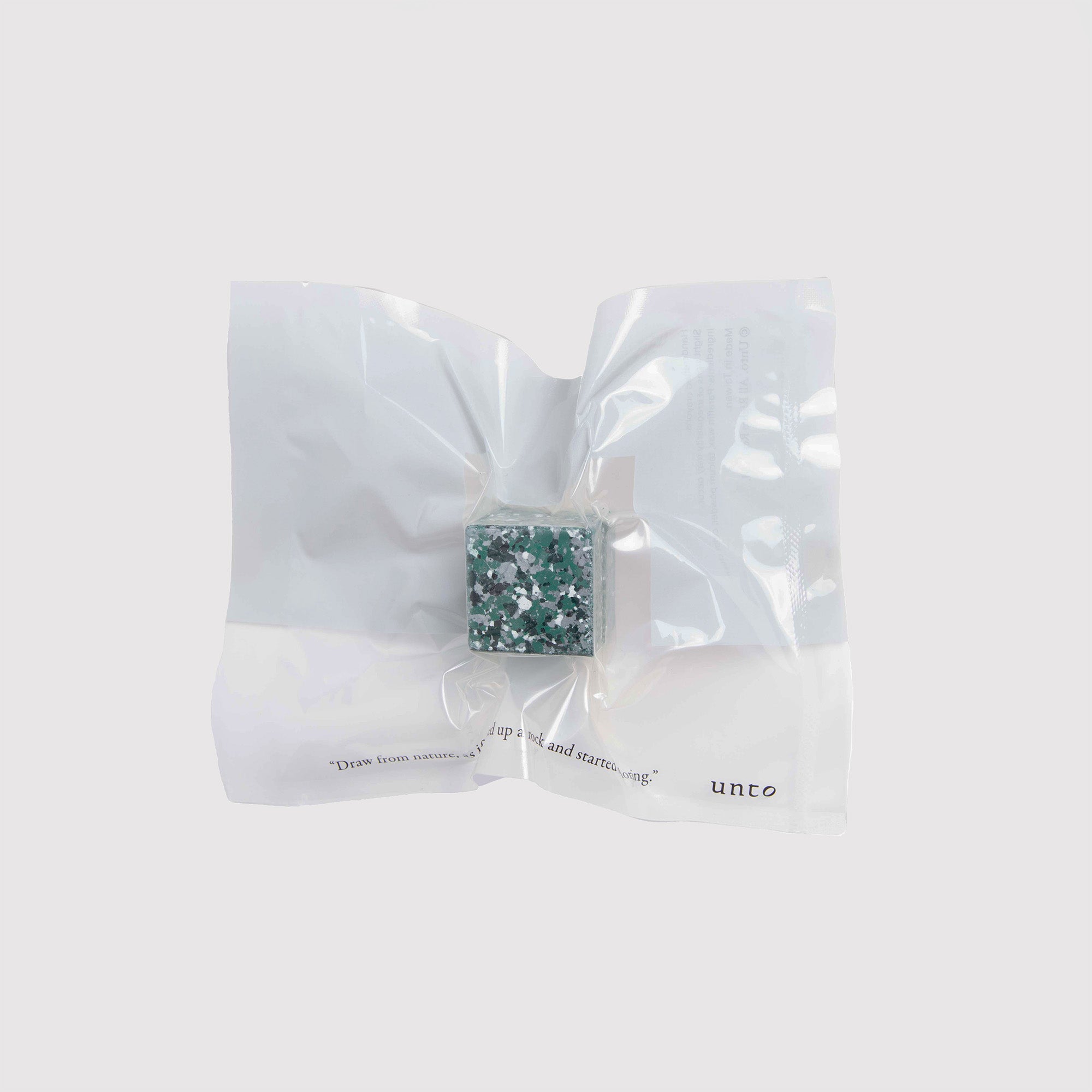 Ein transparenter, vakuumversiegelter Beutel umhüllt einen kleinen quadratischen Block, der einem Color Gem mit grünen und weißen Fragmenten ähnelt. Die minimalistische Verpackung von Studio Untos Color Gem - das Wachsmalquadrat - Einzelpackung zeigt Text in der unteren rechten Ecke auf einem schlichten weißen Hintergrund und strahlt raffinierte Eleganz aus.