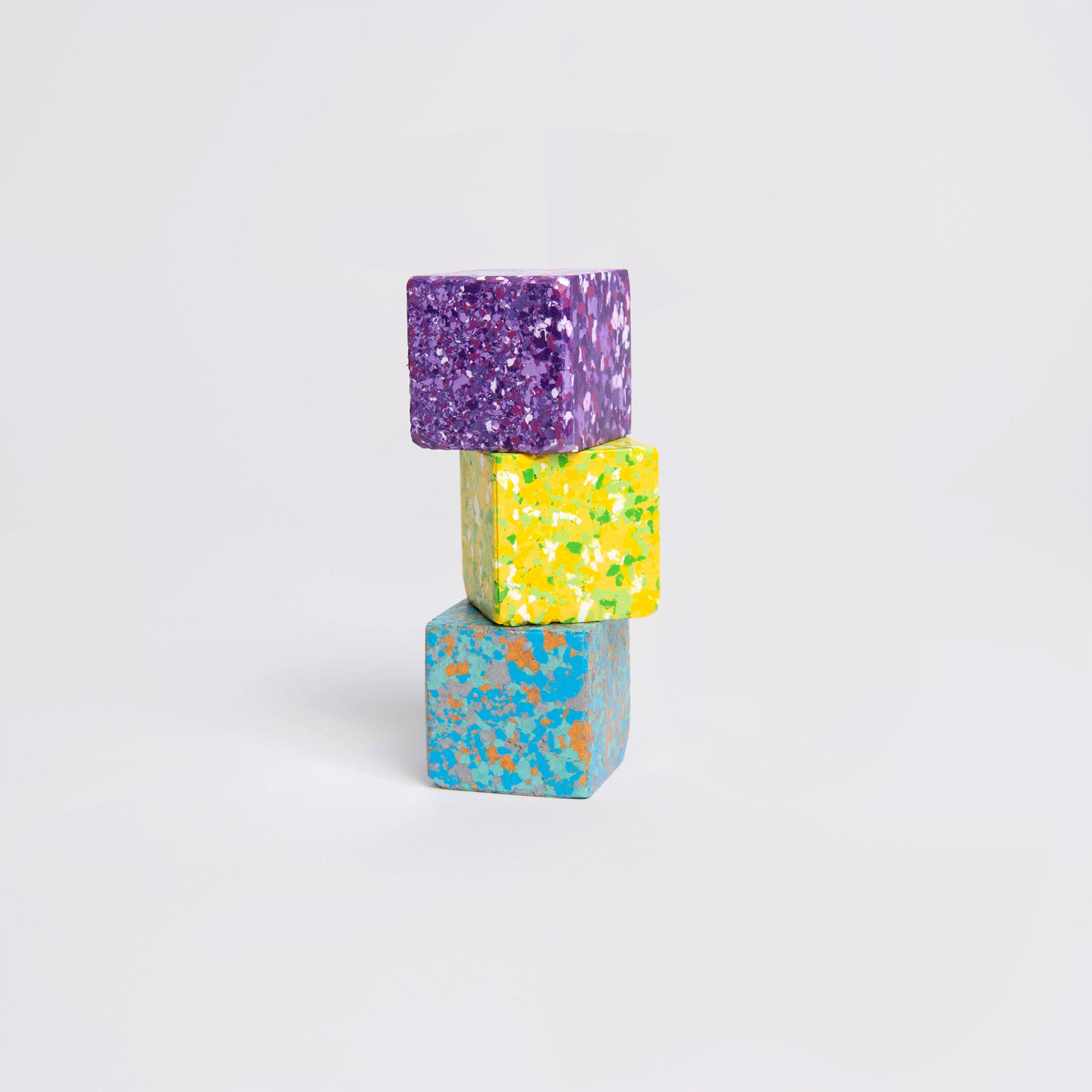 Drei leuchtende Blöcke „Color Gem – das Wachsmalquadrat – Einzelpackung“ von Studio Unto sind vertikal auf einem einfarbigen Hintergrund gestapelt: Der obere Block ist violett mit weißen Sprenkeln, der mittlere Block ist gelb mit bunten Flecken und der untere Block ist blau mit orangefarbenen und grünen Punkten.