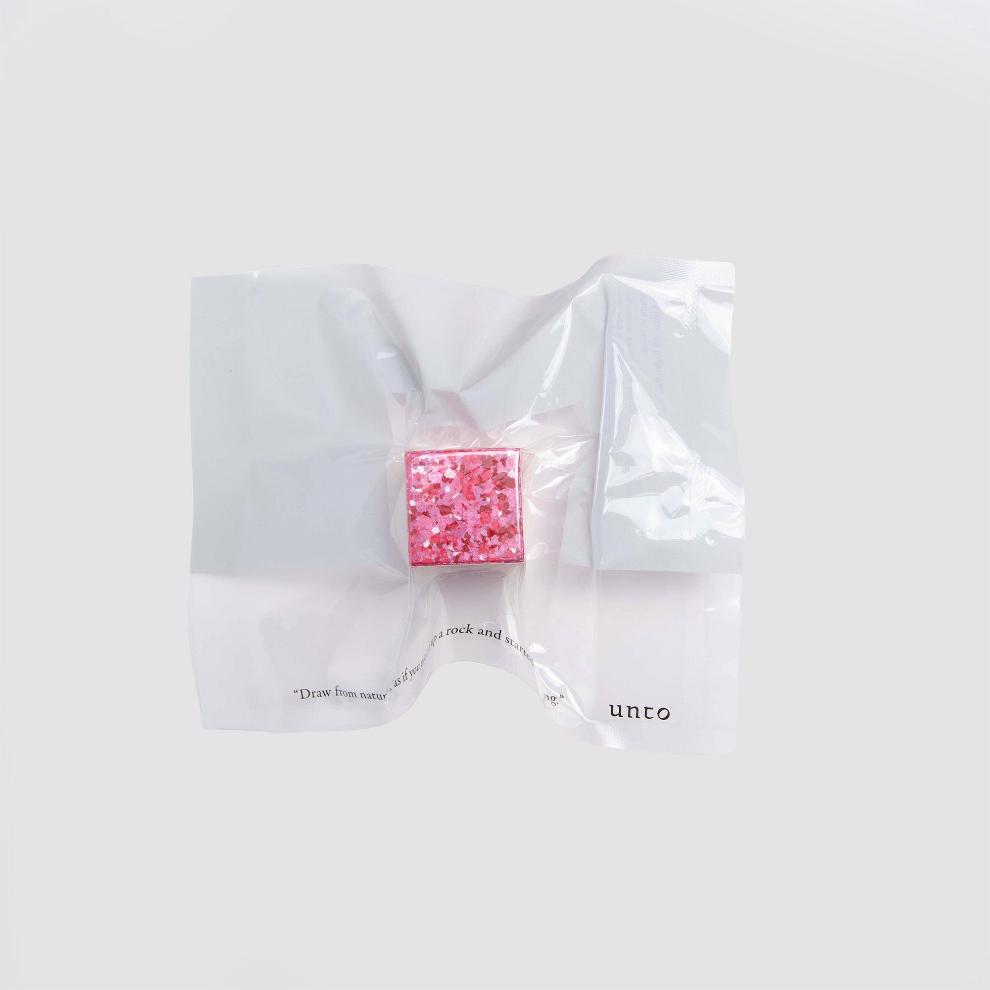 Ein leuchtend rosa Color Gem Würfel mit glitzernder Textur von Studio Unto liegt auf einem zerknüllten weißen Blatt vor hellgrauem Hintergrund und wirkt wie ein dekoratives Accessoire. Er ist Teil der Linie „das Wachsmalquadrat - Einzelpackung“.