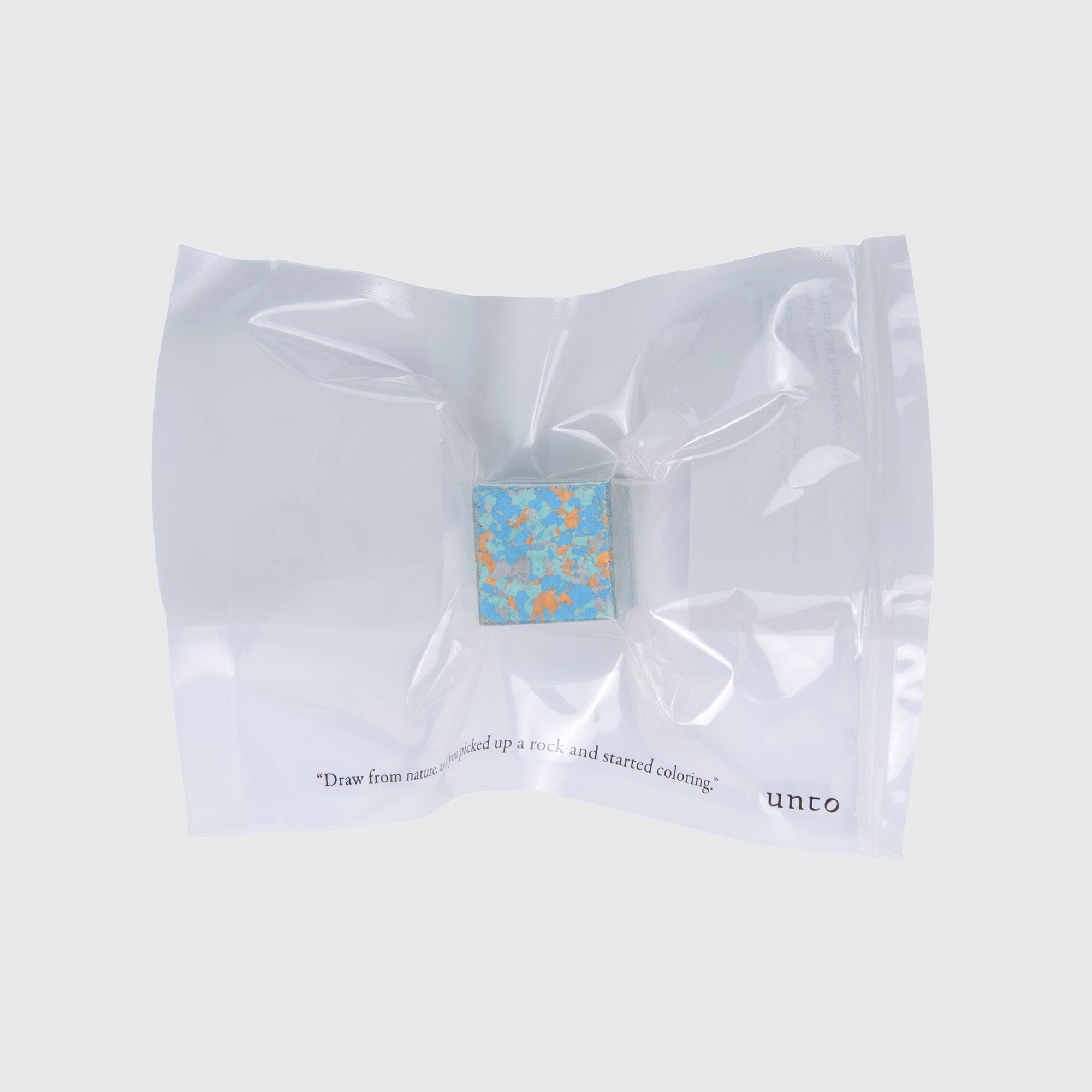 In einem versiegelten transparenten Beutel befindet sich ein kleines, farbenfrohes Color Gem - das Wachsmalquadrat von Studio Unto mit einem marmorierten blau-orangefarbenen Muster. Der Text auf dem Beutel lautet: „Zeichne nach der Natur, nehme einen Stein und beginne zu malen.“ Der Hintergrund ist hellgrau.