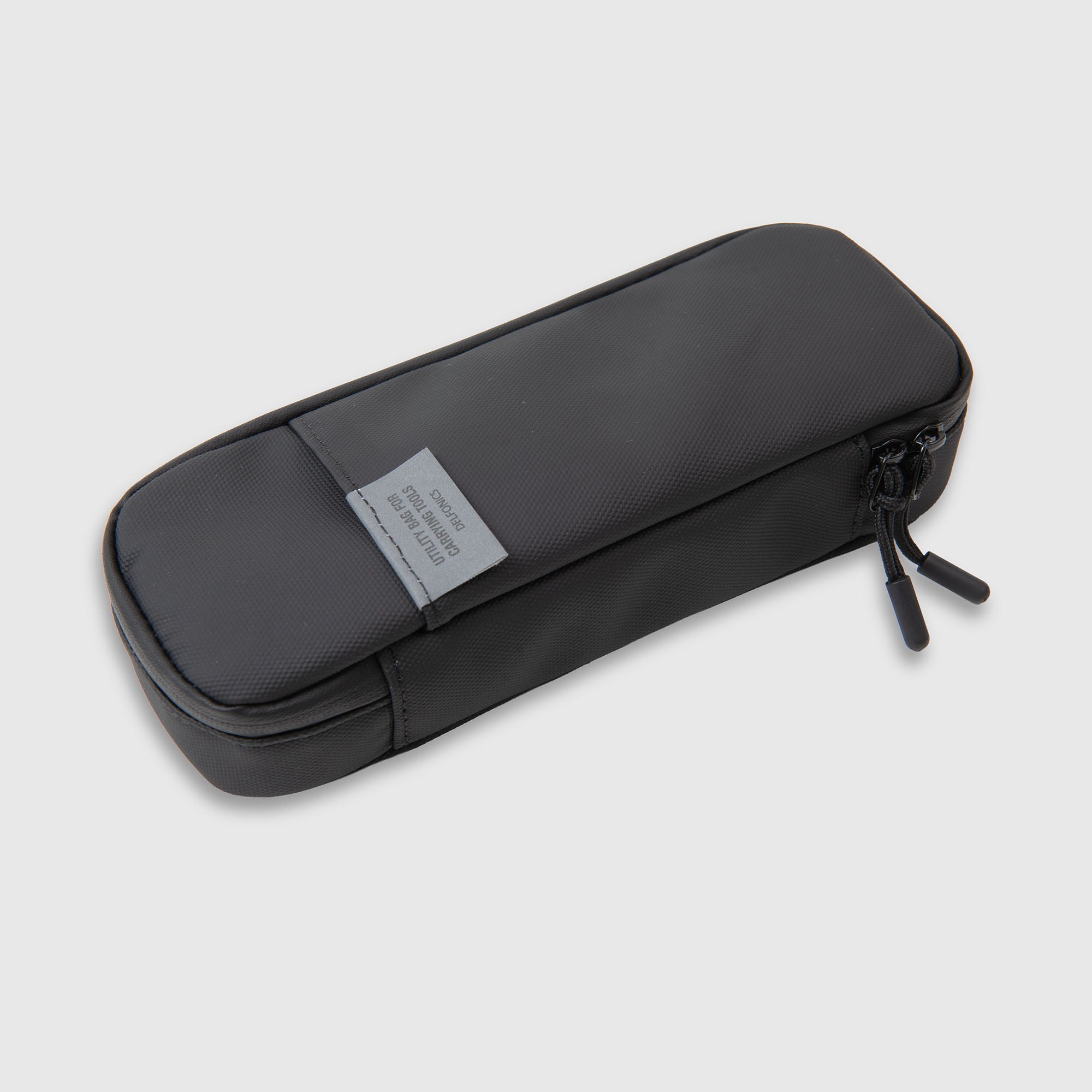 Auf einem hellgrauen Hintergrund ist ein schwarzes, rechteckiges Etui mit Reißverschluss zu sehen: das wasserabweisende „Delfonics Inner Carrying“ Stifteetui von Delfonics mit einem Stoffetikett auf der Oberseite.