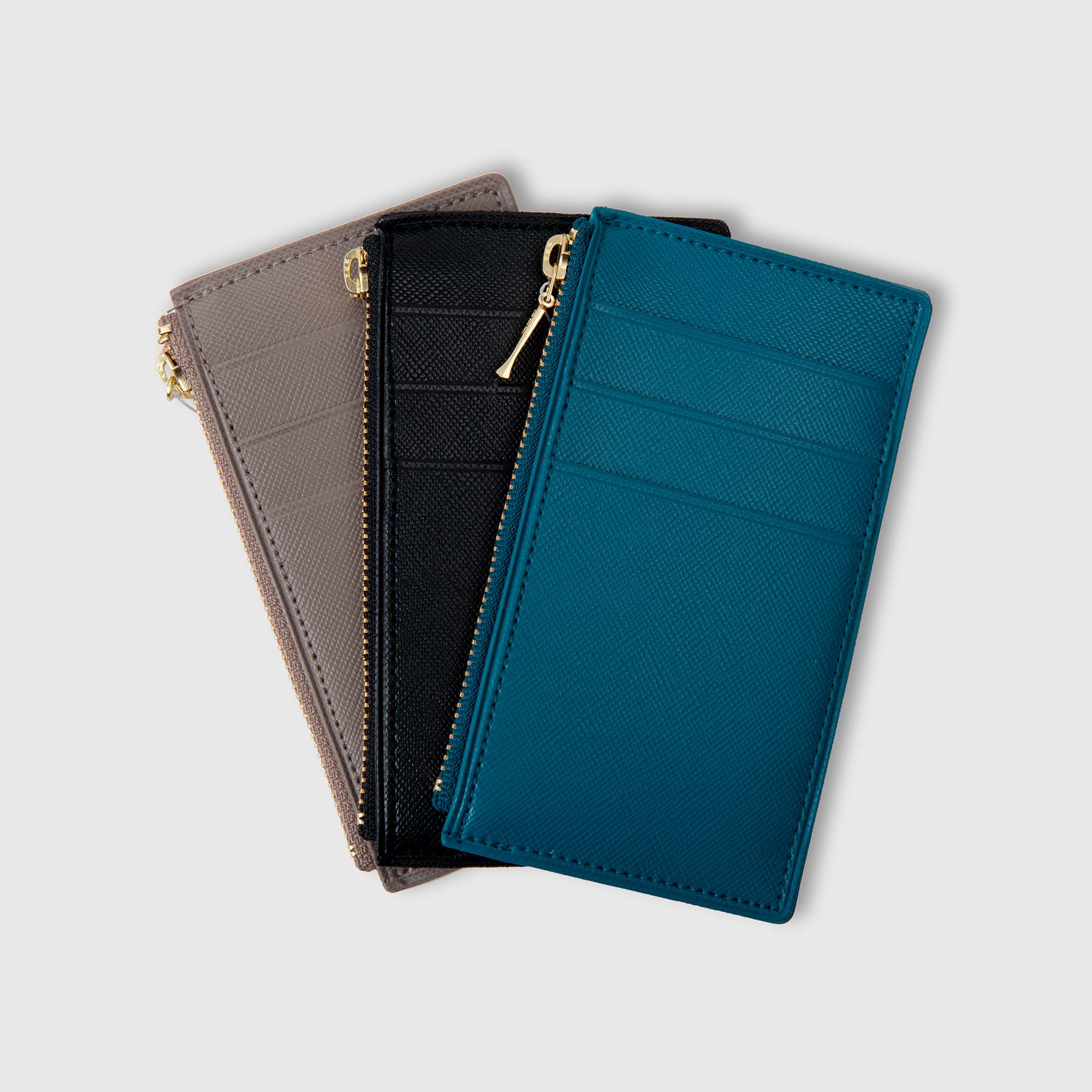Drei schlanke „Quitterie Card Zip Cases“ Kartenetuis von Delfonics in den Farben Taupe, Schwarz und Blaugrün sind auf einem hellgrauen Hintergrund fächerförmig angeordnet. Jedes Etui verfügt über mehrere Kartenfächer und einen goldenen Reißverschluss an der Oberseite.