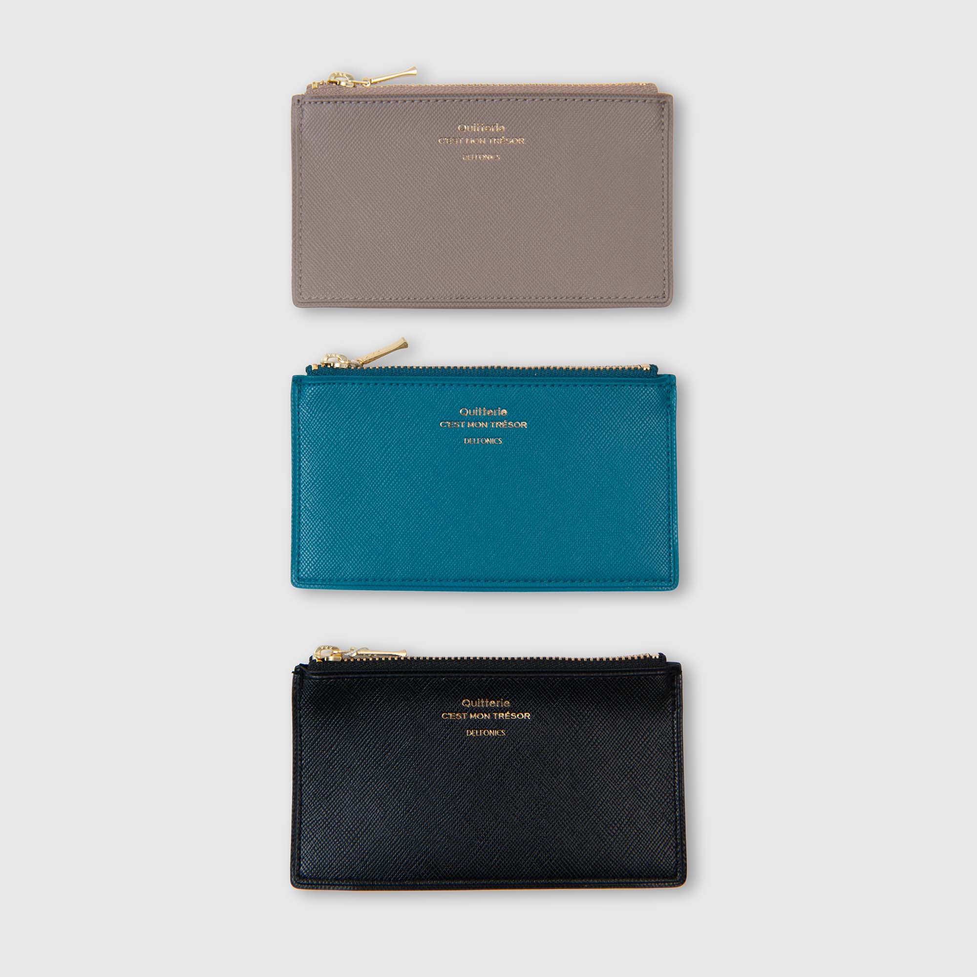 Drei „Quitterie Card Zip Cases“ Kartenetuis von Delfonics – jeweils mit einem goldenen Reißverschluss und goldgeprägten Schriftzügen in den Farben Taupe, Blaugrün und Schwarz – sind vertikal übereinander gestapelt und vor einem hellgrauen Hintergrund ausgestellt.