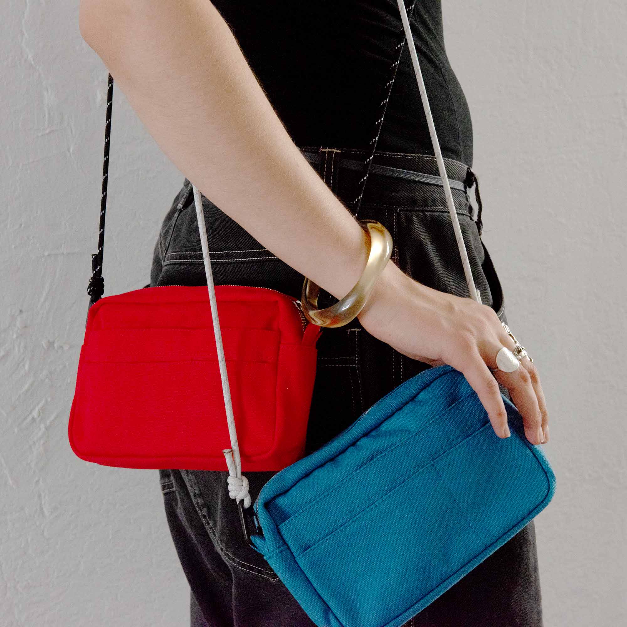 Eine Person, die eine schwarze Hose und ein schwarzes Oberteil trägt, hält eine rote Delfonics-Innentasche und eine blaue Tasche in der Hand, die jeweils mit dem „Delfonics Shoulder Strap Cord for Inner Carrying“ ausgestattet sind – einem verstellbaren Nylon-Schulterriemen von Delfonics. Das Outfit wird durch ein klobiges goldenes Armband und einen silbernen Ring ergänzt.