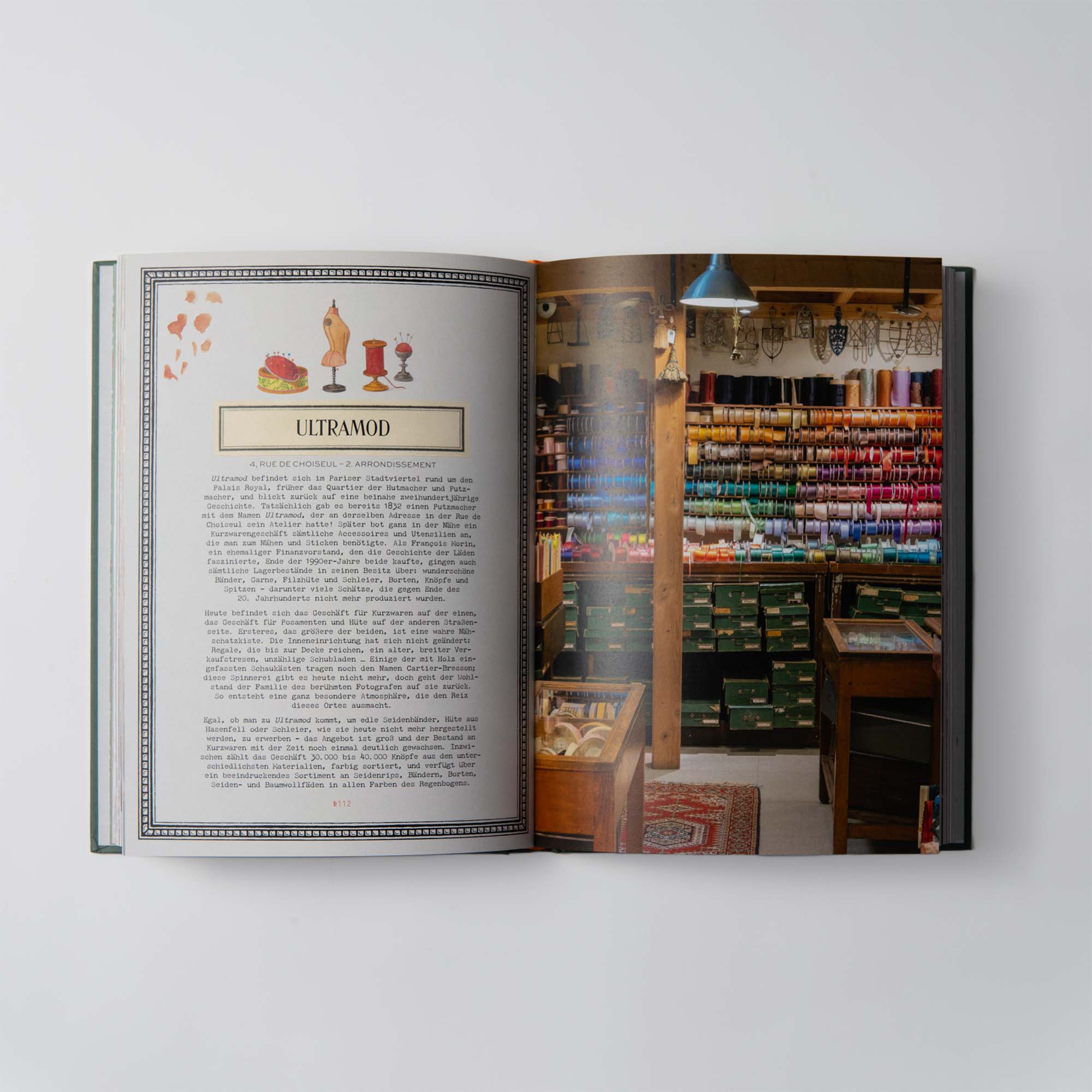 Ein offenes Buch mit dem Titel „Verborgene Schätze in Paris“ vom Dumont Verlag zeigt links ULTRAMOD mit Text und Abbildungen, während rechts ein farbenfrohes Bild eines charmanten Traditionsgeschäfts zu sehen ist. Regale mit farbenfrohen Nähgarnen flankieren einen Holztresen, der Handwerkskunst präsentiert.
