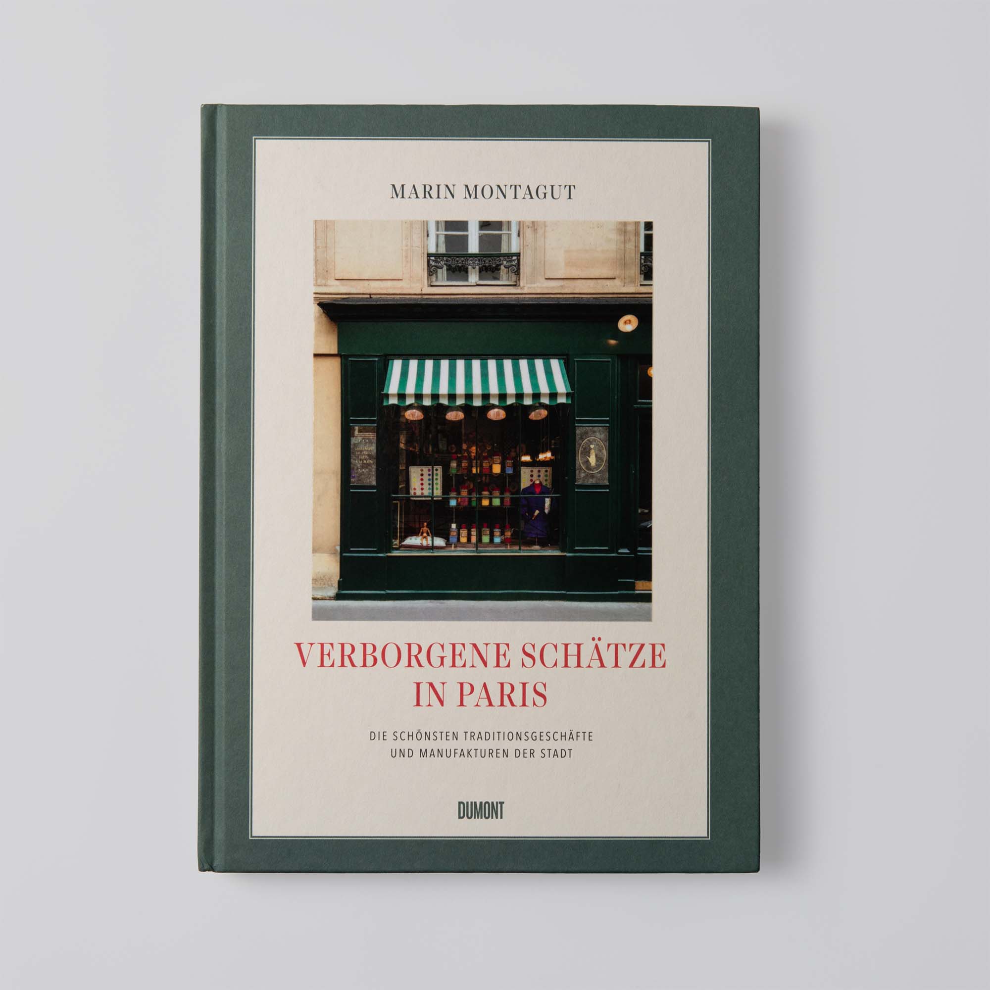 Das Bild zeigt „Verborgene Schätze in Paris“ vom Dumont Verlag, mit einem grünen Einband, der das Foto eines alten Pariser Ladens und einer grün-weiß gestreiften Markise zeigt.