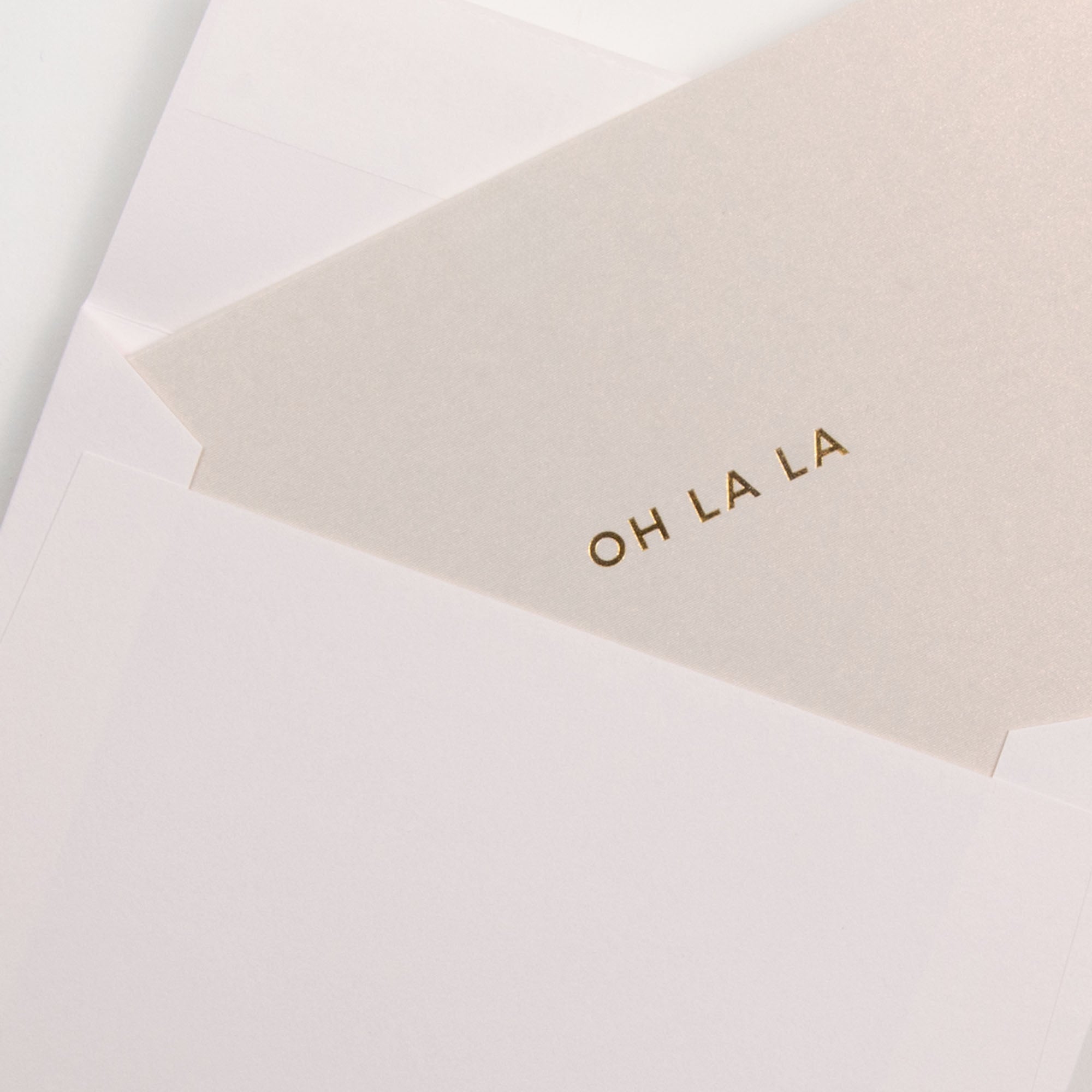 Das Gmunder Kärtchen OH LA LA, eine cremefarbene Letterpress-Karte mit goldener Schrift, wird auf weißem Hintergrund teilweise aus dem passenden Umschlag im A7-Format herausgezogen.