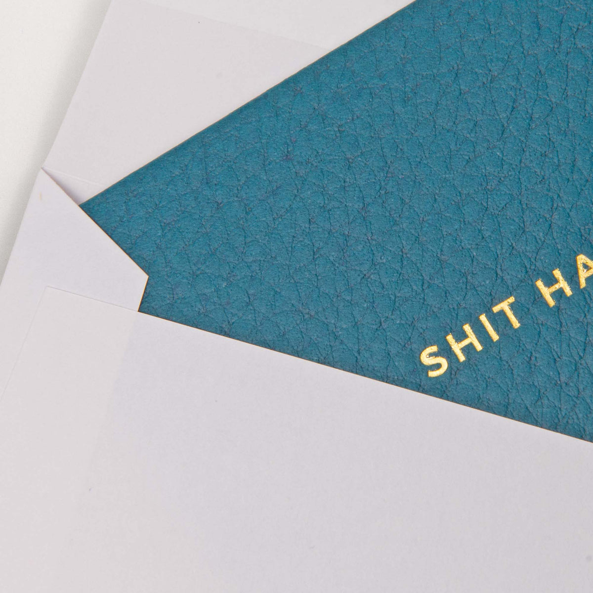 Eine blaue Gmund-Kärtchen-Karte mit der Aufschrift Shit Happens, auf der der Text SHIT HA teilweise zu sehen ist, in einem weißen Umschlag.