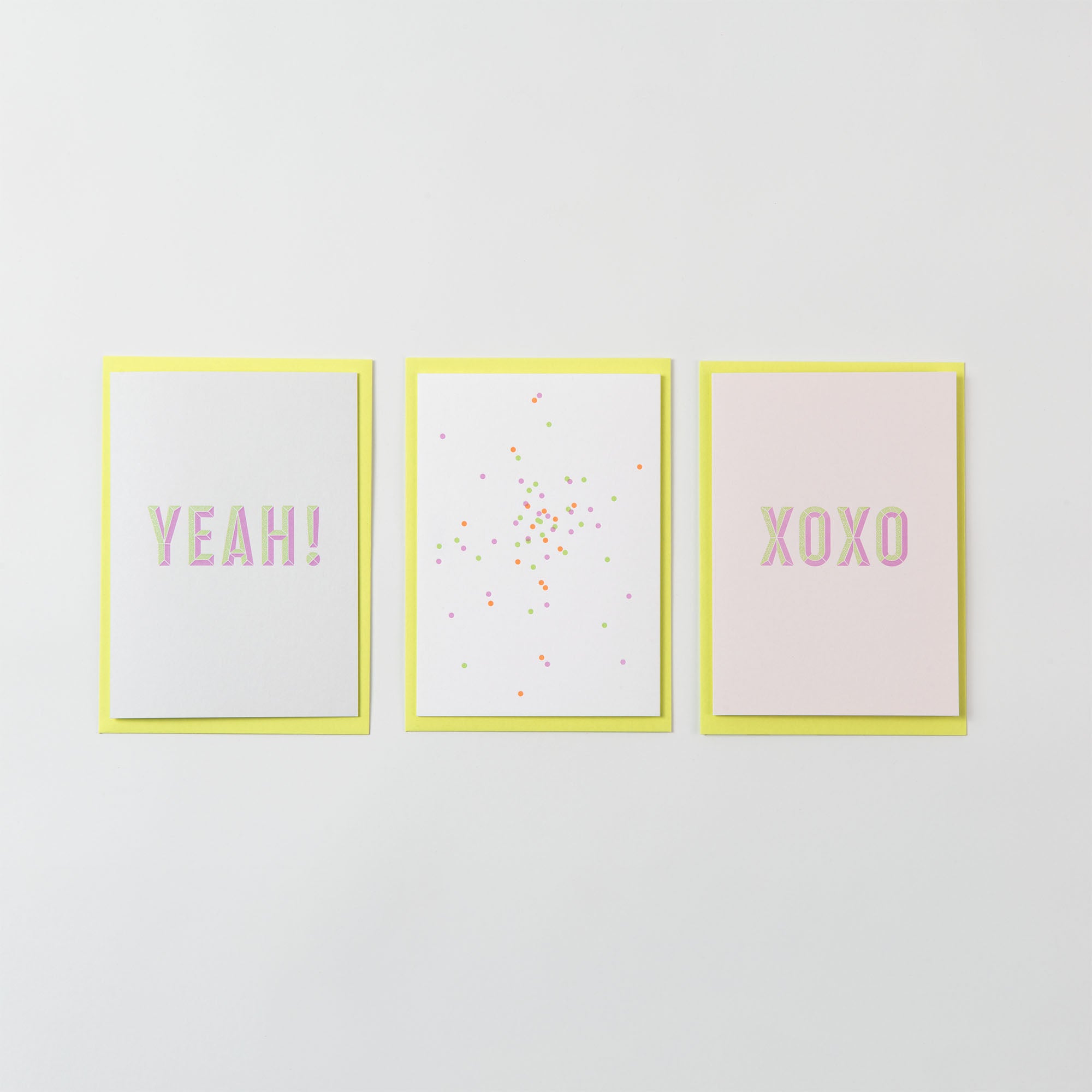 Das Grußkartenset YEAH & Konfetti von paperlux.store besteht aus drei nebeneinander angeordneten Grußkarten mit jeweils blassgelben Rändern. Auf der ersten steht „YEAH!“, auf der mittleren sind bunte Punkte zu sehen und auf der dritten steht in rosa Buchstaben „XOXO“. Alle Karten sind im Letterpress-Verfahren aus Gmund-Papier gefertigt.