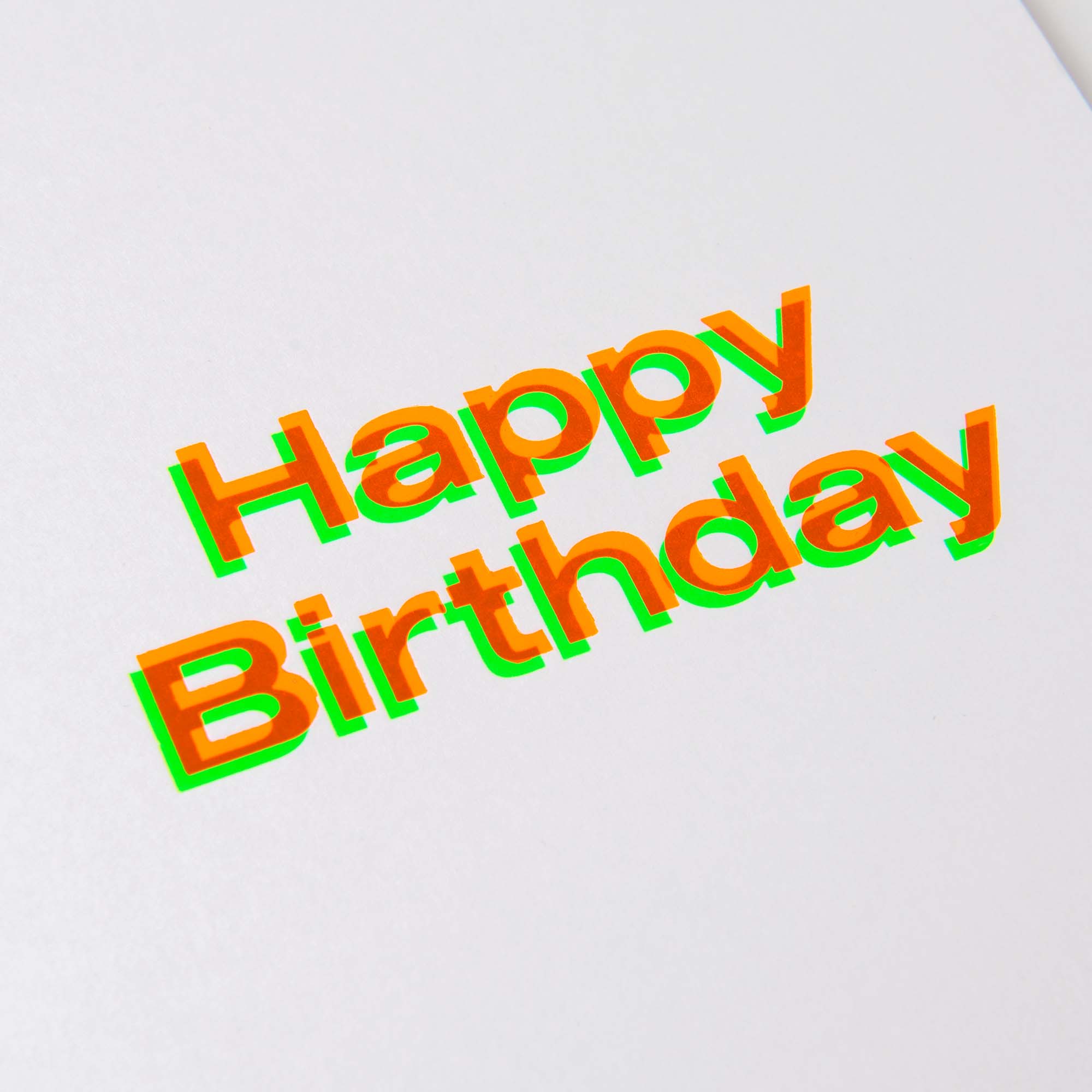 Die „Happy Birthday Karte A5“ von paperlux.store zeigt einen fettgedruckten, überlappenden orangefarbenen Text mit einem schimmernden grünen Schatten und erinnert so an traditionelle Geburtstagskarten. Das elegante Design wirkt wie in Hamburg gefertigt, vor einem schlichten weißen Hintergrund.