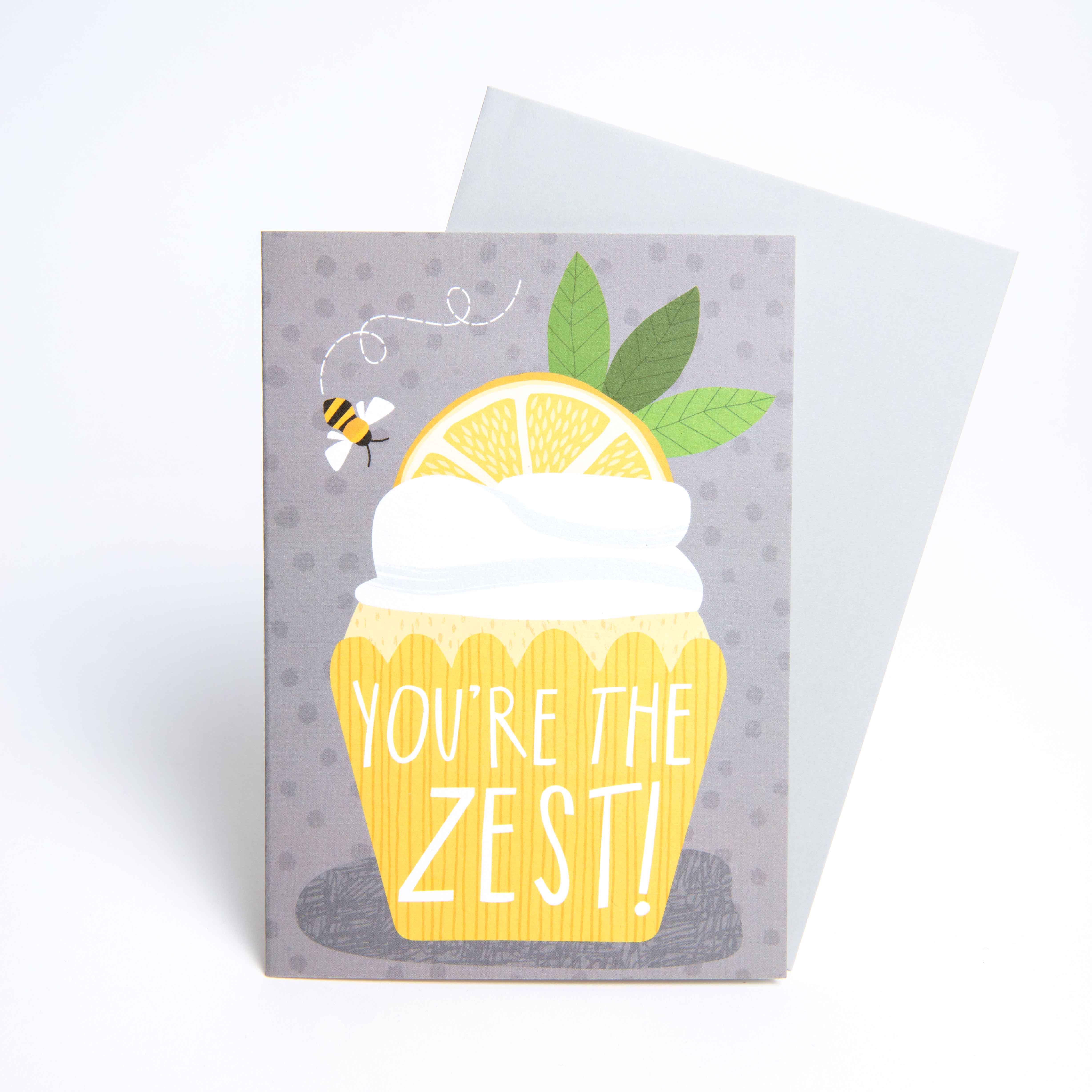 Die Grußkarte Lemon von Joanne de Pace zeigt einen gelben Cupcake mit weißem Zuckerguss, einer Zitronenscheibe und Blättern. In der Nähe schwebt eine Biene mit dem Text Youre the Zest! vor einem grau gepunkteten Hintergrund. Ein Umschlag für all Ihre Grußkarten-Bedürfnisse ist im Lieferumfang enthalten.