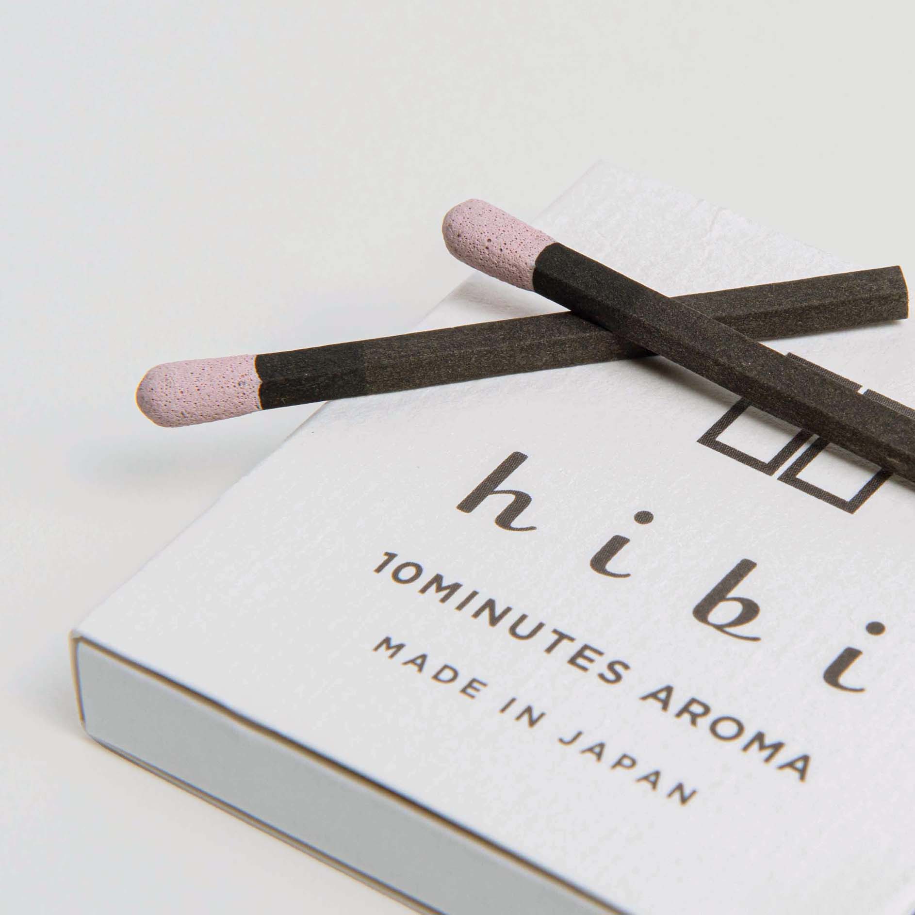 Zwei Räucherstäbchen mit rosa Spitze liegen auf einer teilweise sichtbaren weißen Schachtel mit der Aufschrift „hibi 10 Minuten Aroma, hergestellt in Japan“. Die Duftstreichhölzer von Hibi sorgen für ein natürliches Dufterlebnis auf schlichtem weißen Hintergrund.