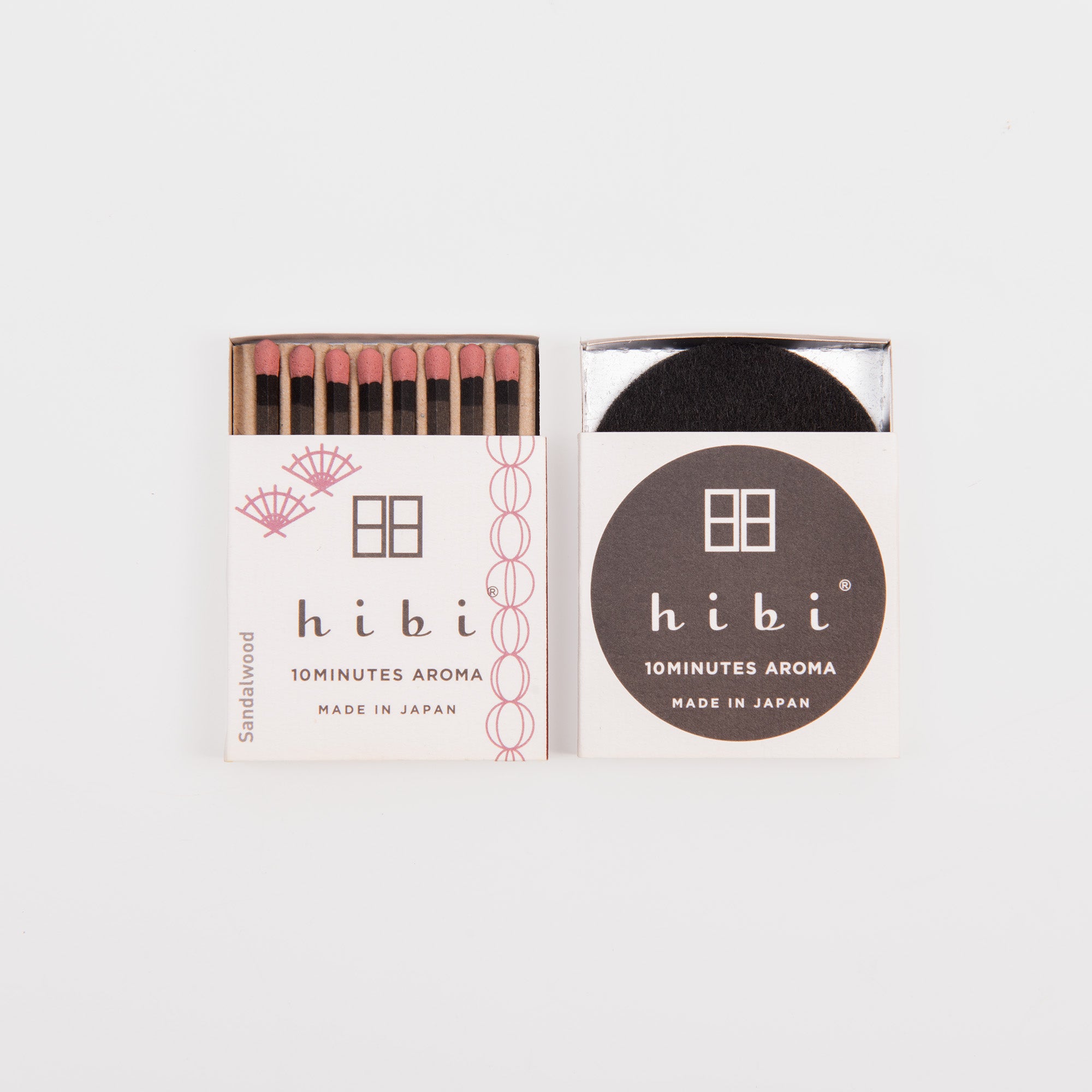 Zwei geöffnete Hibi - Duftstreichhölzer liegen auf einer weißen Streichholzschachtel, die eine zeigt rosafarbene Sandelholzstreichhölzer, die andere das Hibi-Logo, "10 MINUTES AROMA MADE IN JAPAN" und einen runden schwarzen Block, ideal zur Entspannung.