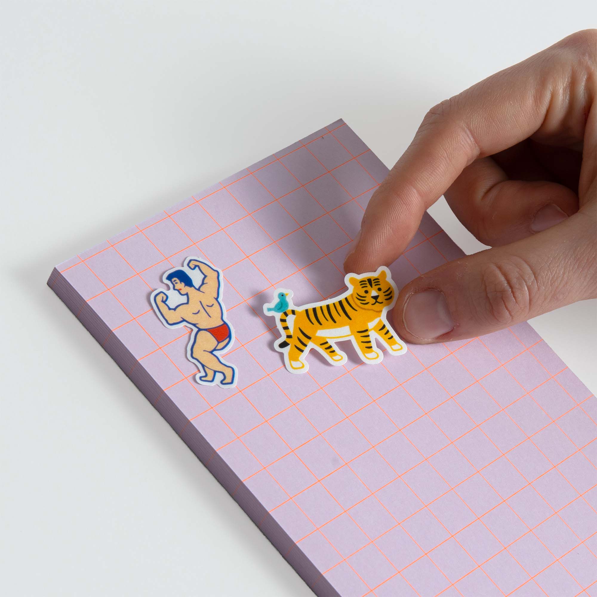 Eine Hand platziert einen Hightide Flake Sticker von Hightide - einen Tiger mit einem Vogel auf dem Rücken - auf einem rosa Notizblock mit einem roten Gitter. Ein weiterer Aufkleber aus der Serie Laidback Animals, eine sich windende Figur in blauen Shorts, befindet sich bereits auf dem Notizblock.