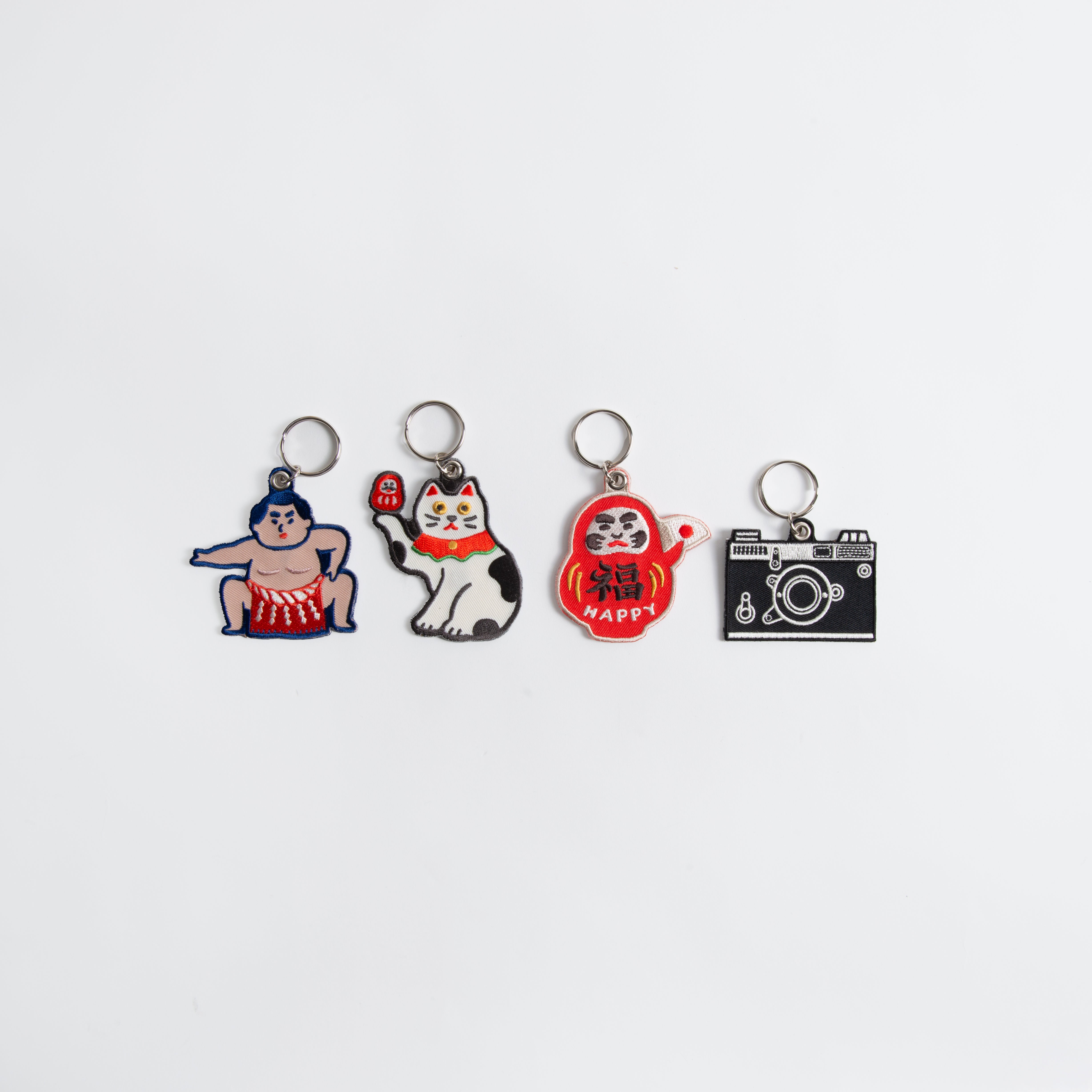Vier Hightide Stitch Work Schlüsselanhänger von Hightide-Kamera, Daruma & Glückskatze auf weißem Hintergrund: perfekte japanische Geschenkideen oder bestickte Schlüsselanhänger für Sammler.