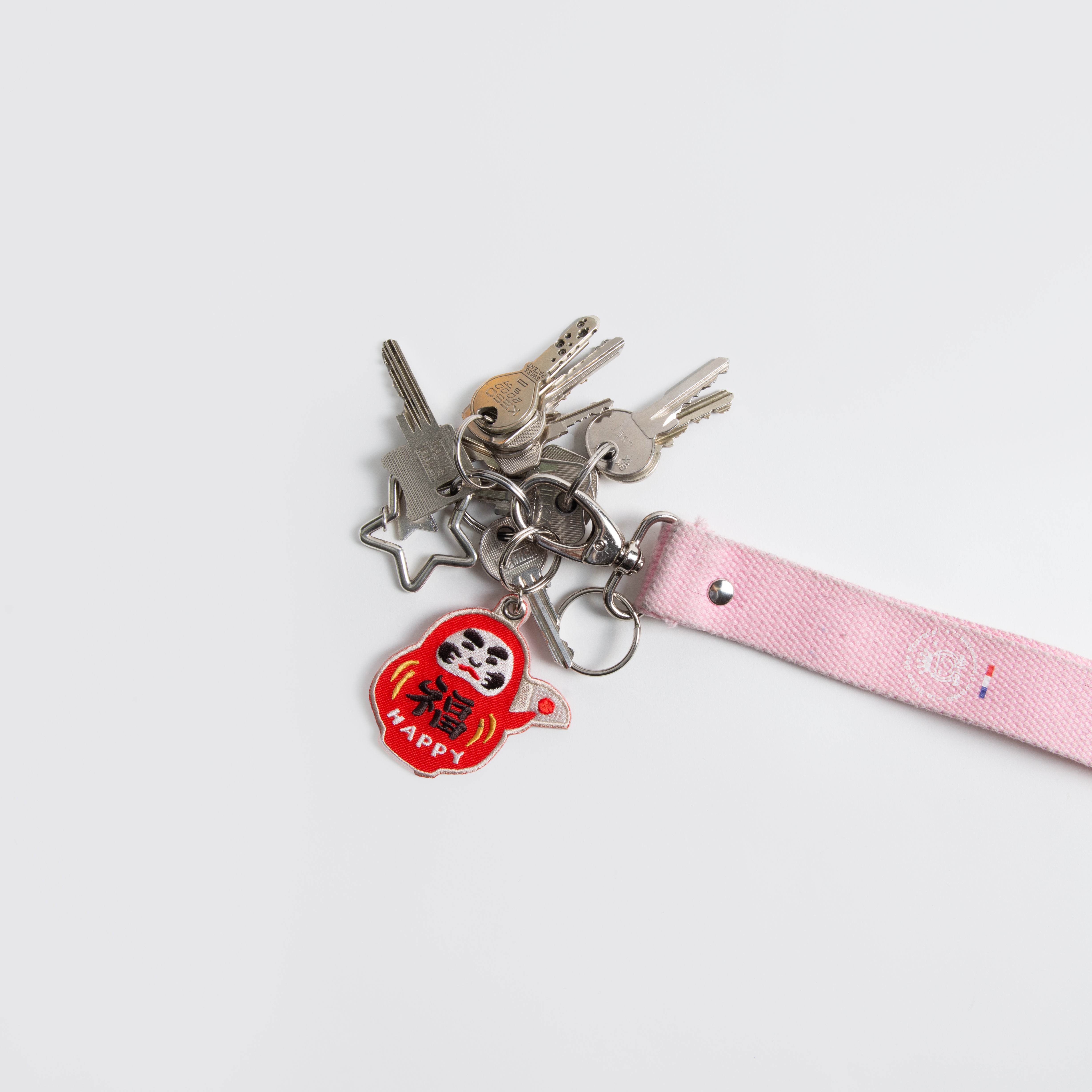 Ein Set von Hightide Stitch Work Schlüsselanhängern - Kamera, Daruma & Glückskatze - von Hightide, bestehend aus einem rosa Band, einem silbernen sternförmigen Schlüsselanhänger und einem roten Daruma "HAPPY" Anhänger; ideal für japanische Geschenkideen auf weißem Hintergrund.