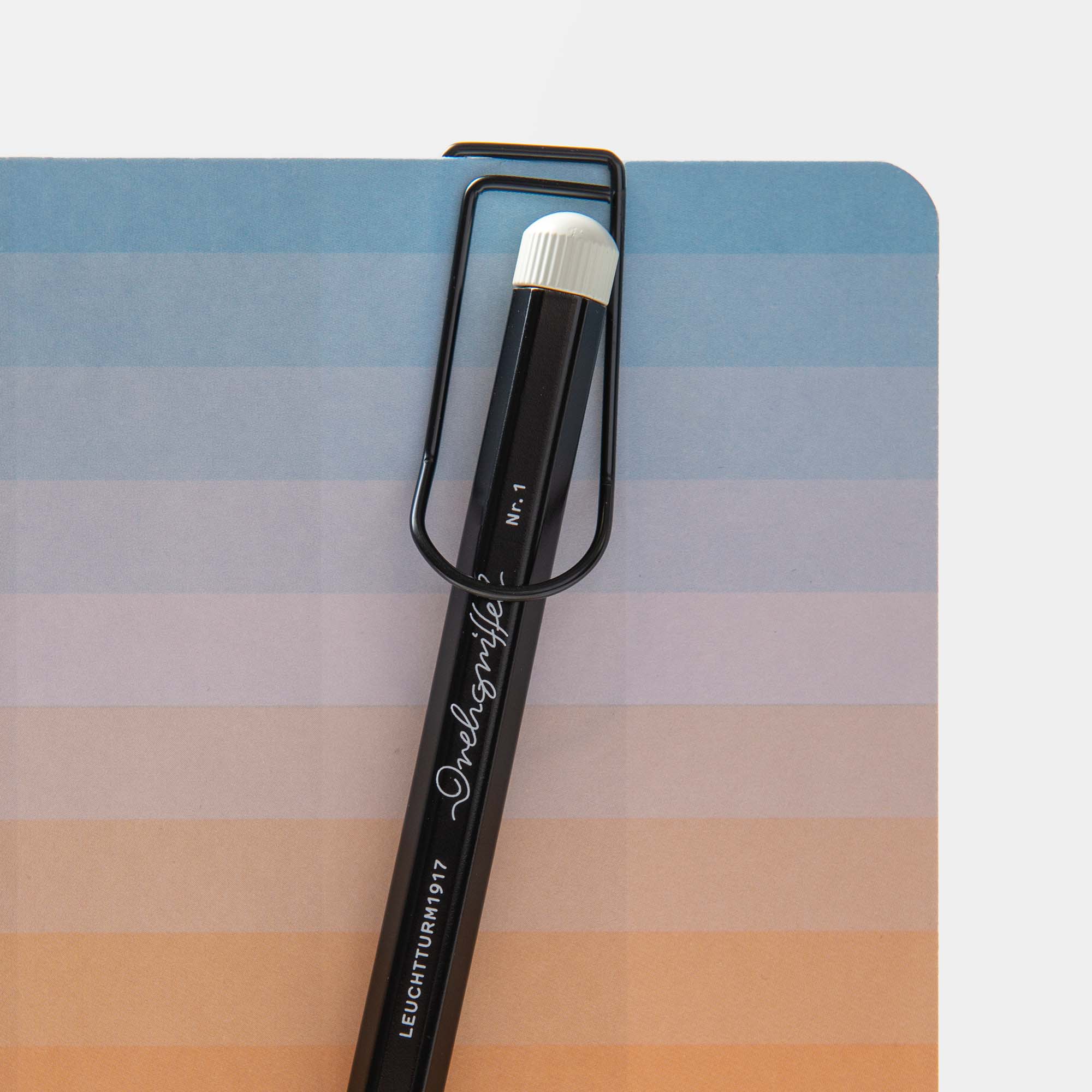 Ein großer, schwarzer Hightide Pen Hook Clip - Stiftehalter To Go sichert einen schwarzen Bleistift, der einem eleganten Kugelschreiber ähnelt, mit eleganter weißer Schrift und einem weißen Radiergummi vor einem blau und orange gestreiften Notizbuchumschlag.