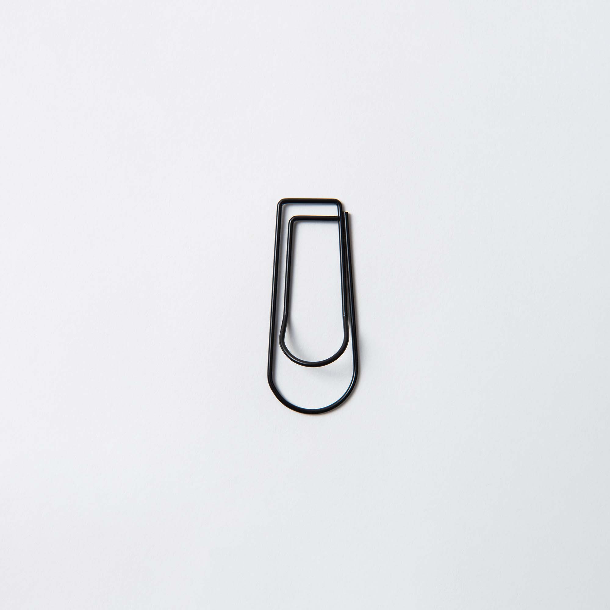 Der Pen Hook Clip - Stiftehalter To Go von Hightide ist ein einfacher schwarzer Clip, der an eine Eisenklammer erinnert und auf weißem Hintergrund angebracht ist. Sein minimalistisches Design zeigt eine glatte, längliche ovale Form.