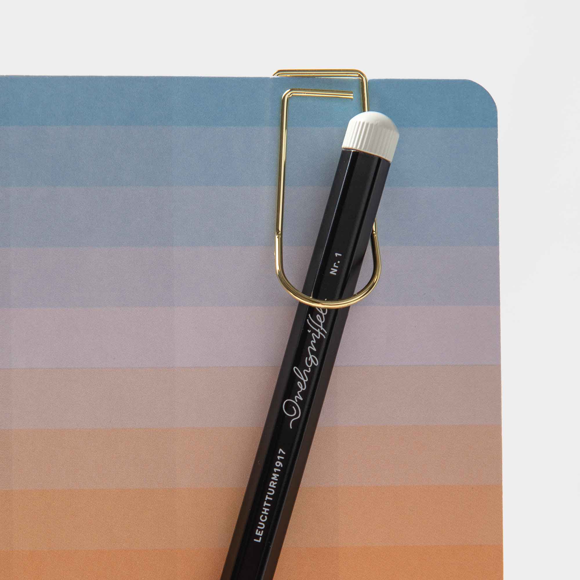 Ein schwarzer Bleistift mit weißem Radiergummi wird mit dem Pen Hook Clip – Stiftehalter To Go von Hightide an einem Notizbuch befestigt. Das Notizbuch hat einen Farbverlaufs-Umschlag in Blau und Orange, der an bunte Notizbücher erinnert.