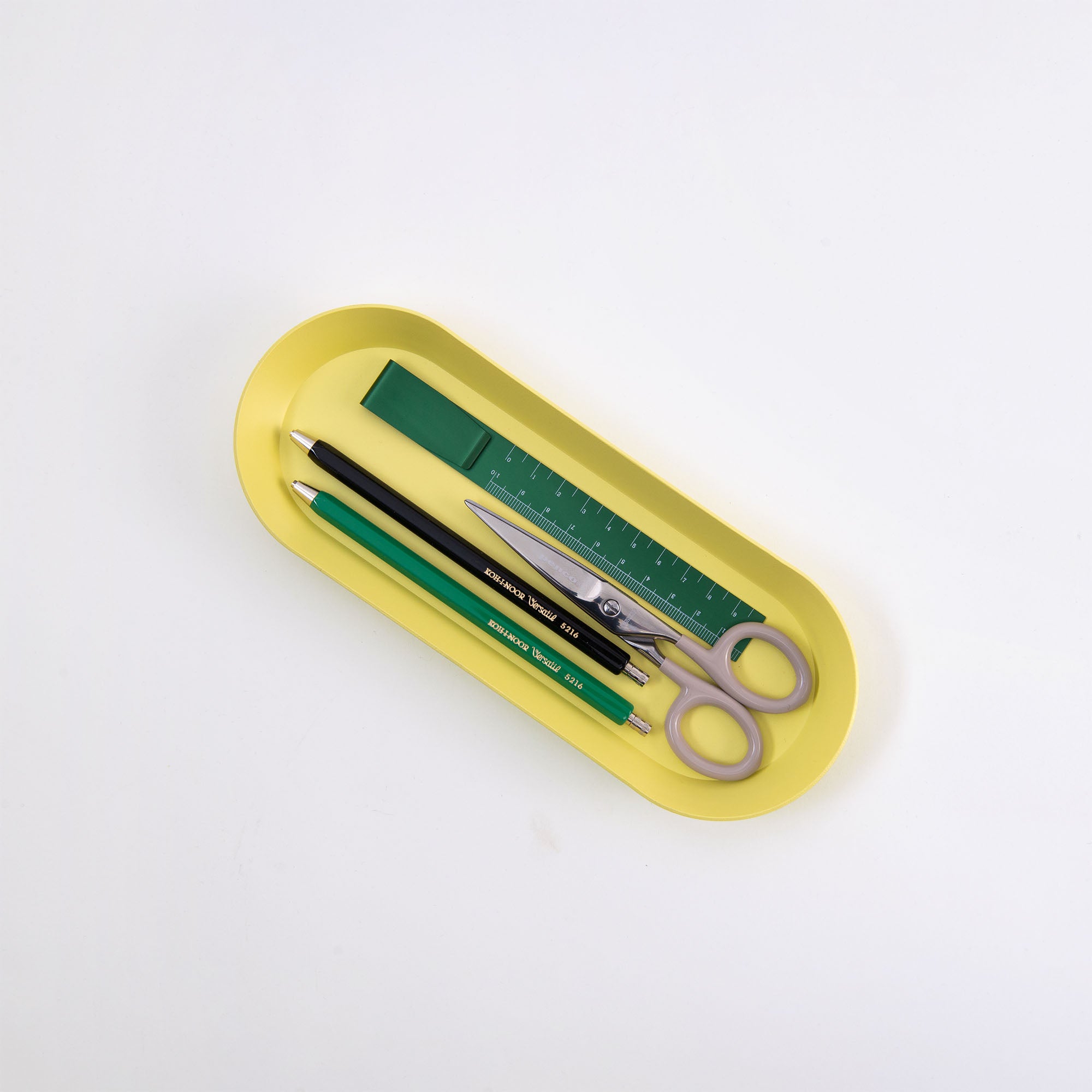Das gelbe Tablett „Ovale Schale“ von Hightide verkörpert mit seinen ordentlich angeordneten Schreibwaren ein Musterbeispiel für Ordnung: ein grünes Lineal, eine Schere mit grauem Griff, ein Druckbleistift, ein schwarzer Stift und ein grünes Radiergummi.