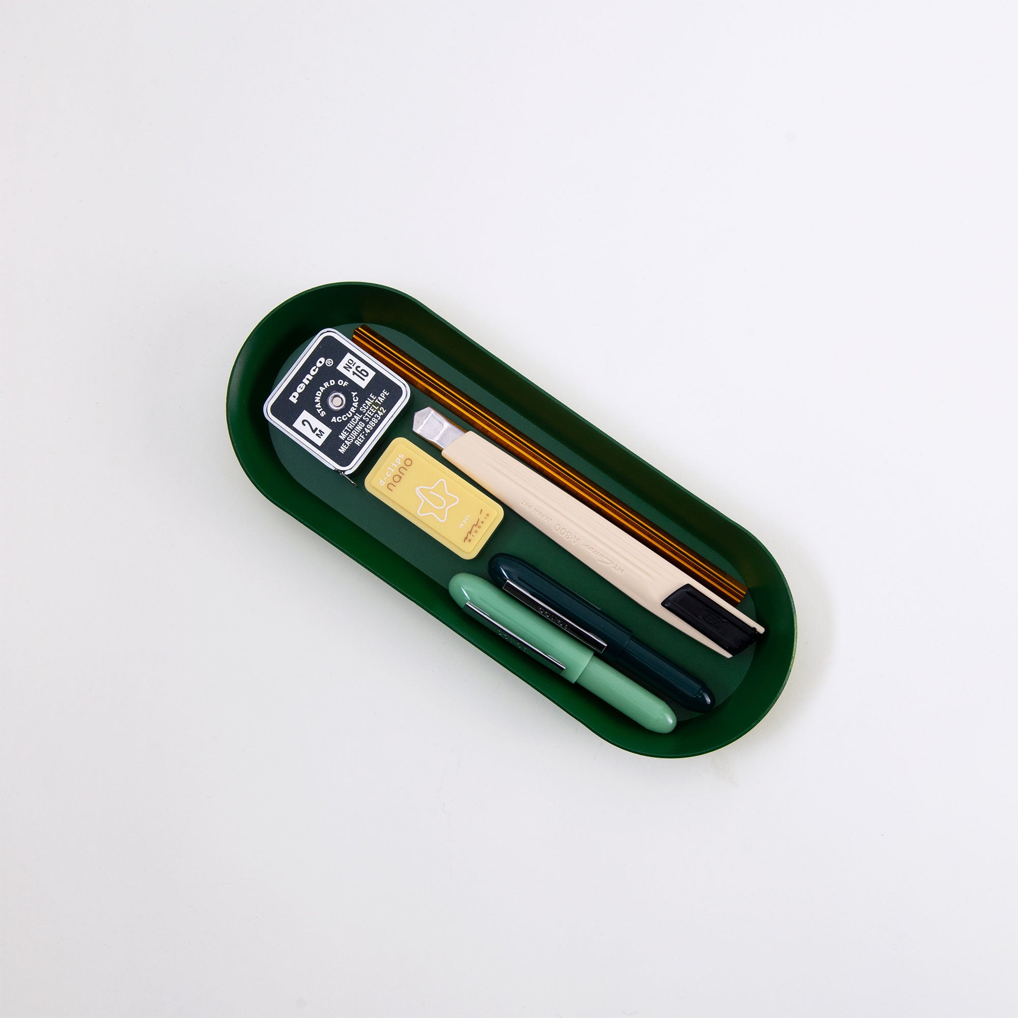 Die grüne ovale Schale von Hightide enthält einen Bleistift, einen Stift, einen gelben Radiergummi mit Sterndesign, einen schwarzen Anspitzer mit weißem Text und ein Holzlineal. Alle Artikel sind ordentlich angeordnet.