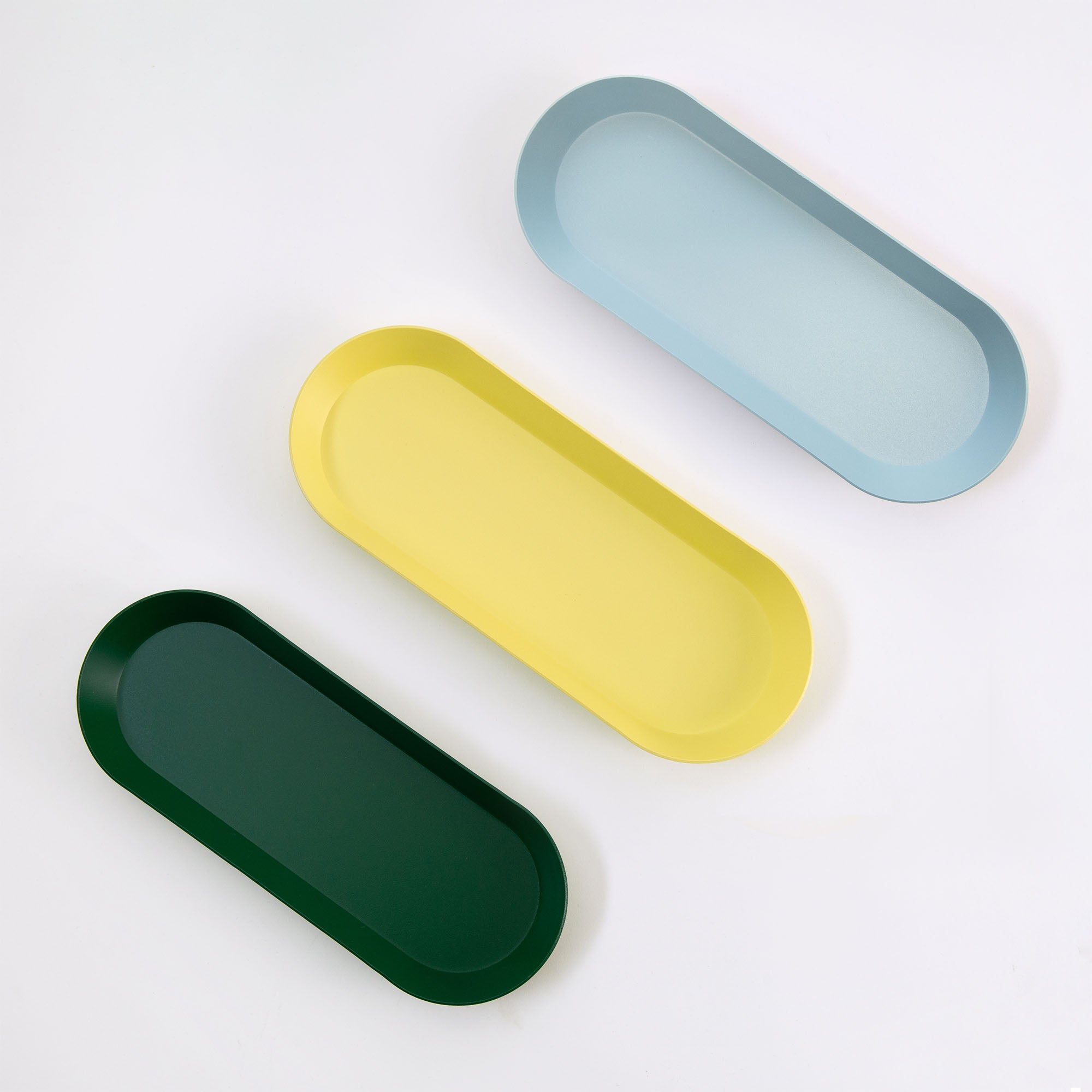 Drei Tabletts der Ovalen Schale von Hightide sind diagonal auf weißem Hintergrund im Ordnungsstil angeordnet. Das minimalistische Design zeigt Tabletts in Dunkelgrün, Gelb und Hellblau von unten nach oben.