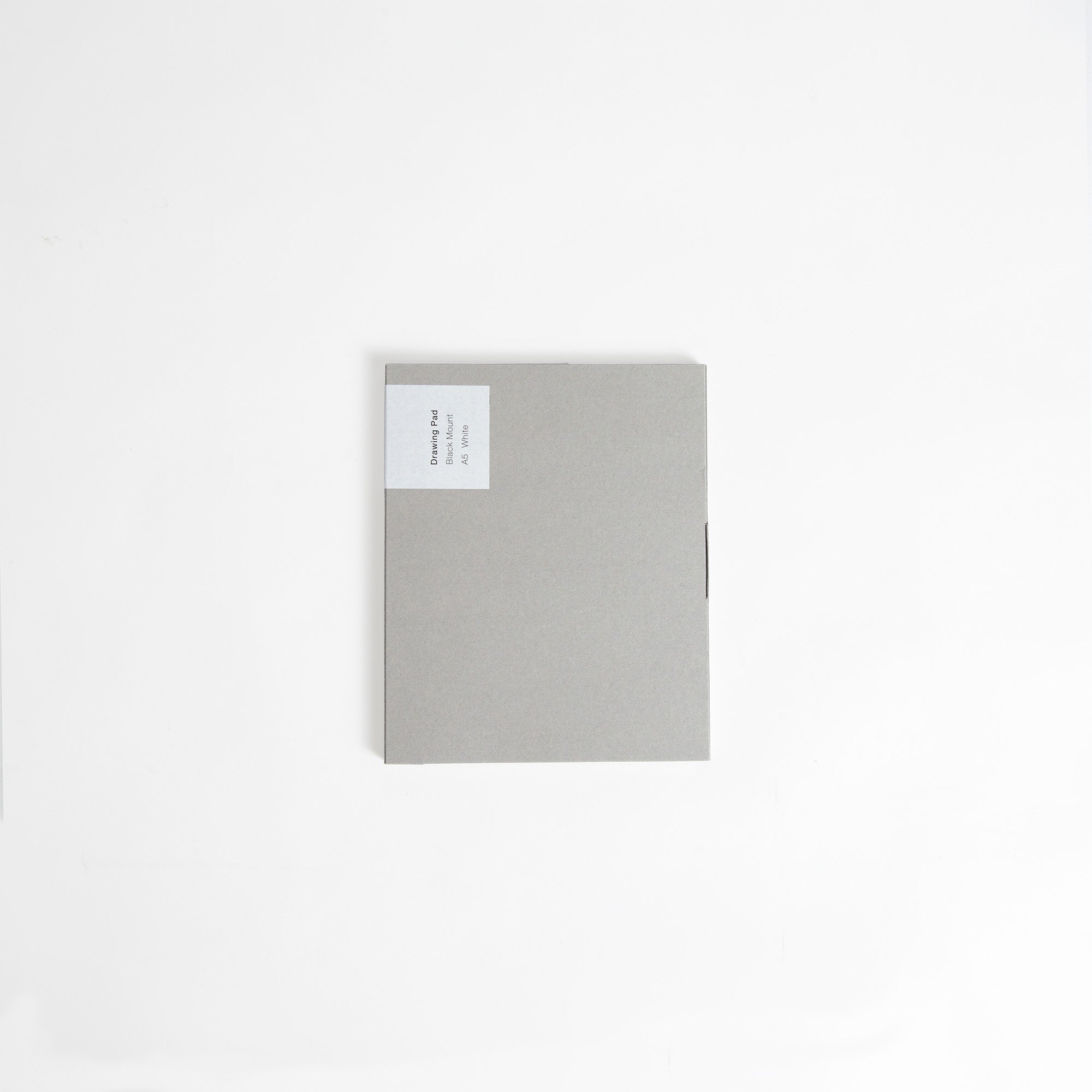 Ein quadratischer, hellgrauer ITO Bindery Drawing Pad Black Mount von ITO Bindery liegt geschlossen auf einer weißen Fläche. Ein kleines weißes Etikett mit aufgedrucktem Text befindet sich in der oberen linken Ecke.