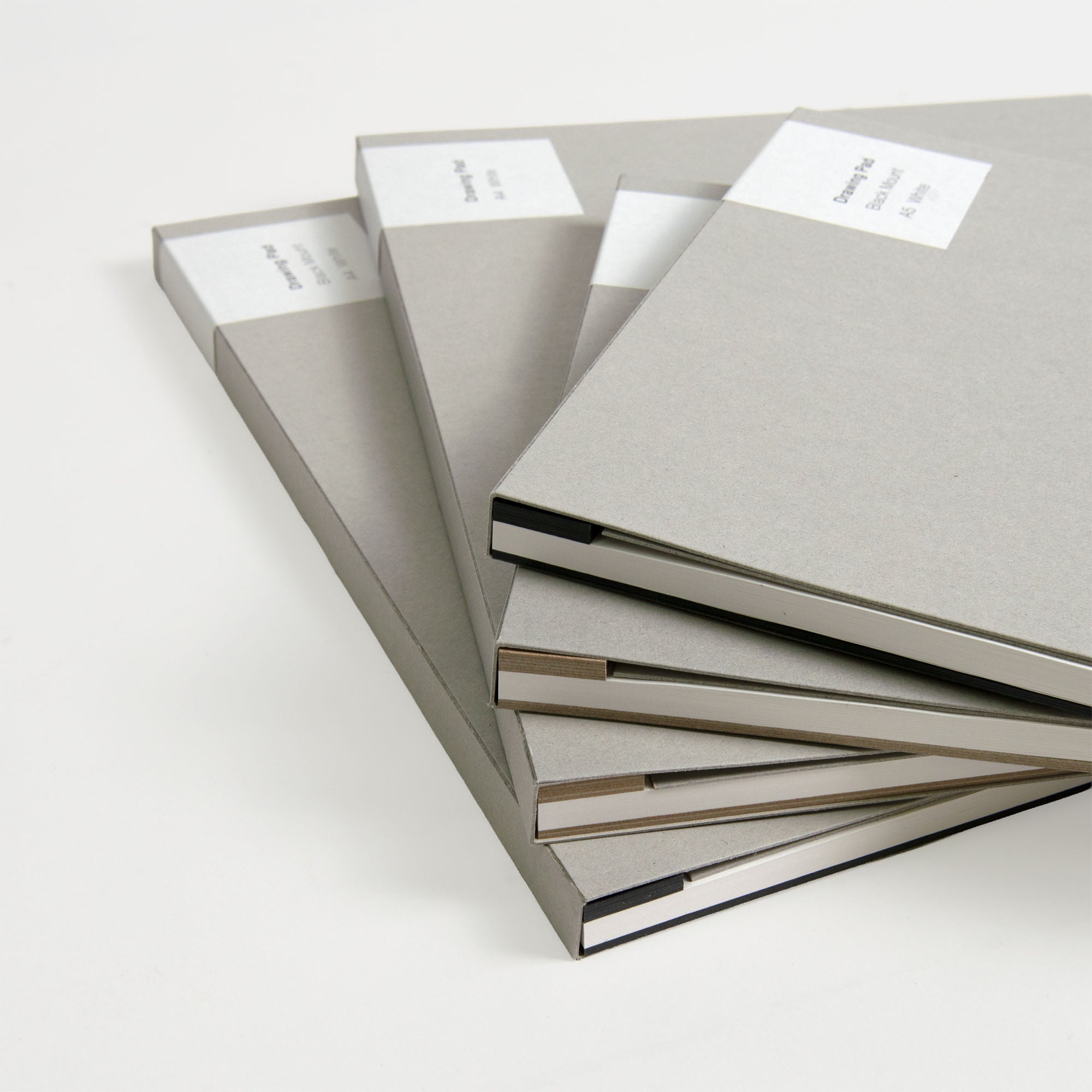 Vier ITO Bindery Drawing Pad White Bücher von ITO Bindery sind ordentlich und leicht aufgefächert auf einer weißen Fläche gestapelt. Sie präsentieren ihre grauen Hardcover mit weißen rechteckigen Etiketten und einer Eleganz, die an japanisches Papier erinnert.