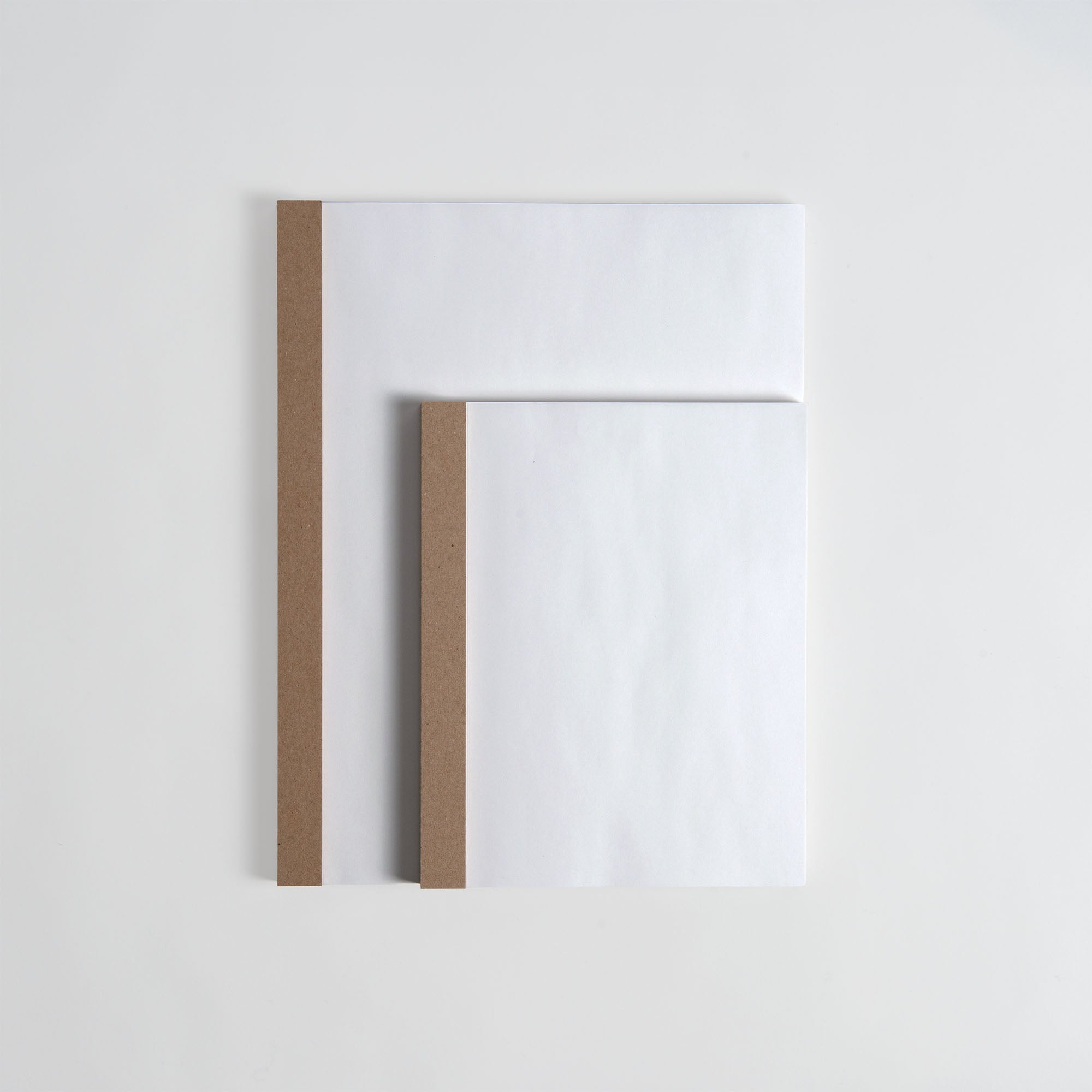 Zwei minimalistische ITO Bindery Drawing Pad White Notizbücher - ein großes und ein kleines - liegen überlappend auf einer weißen Fläche. Ihre braunen Buchrücken und leeren Einbände strahlen Schlichtheit und Eleganz aus. Hergestellt von ITO Bindery mit feinem Japanpapier.