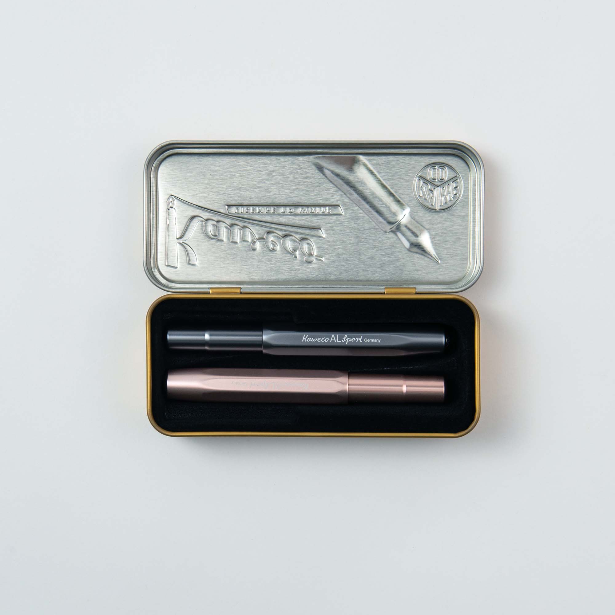 Die Blechdose mit dem Markennamen „Kaweco“ und Aluminiumgehäuse besticht durch ein erhabenes Füllfederhalter-Design auf dem Deckel. Im Inneren befindet sich ein klassisches schwarz-silbernes Füllhalter M-Stifteset in einem gefütterten Fach. Der kompakte Deckel ziert außerdem ein geprägtes rundes Emblem.