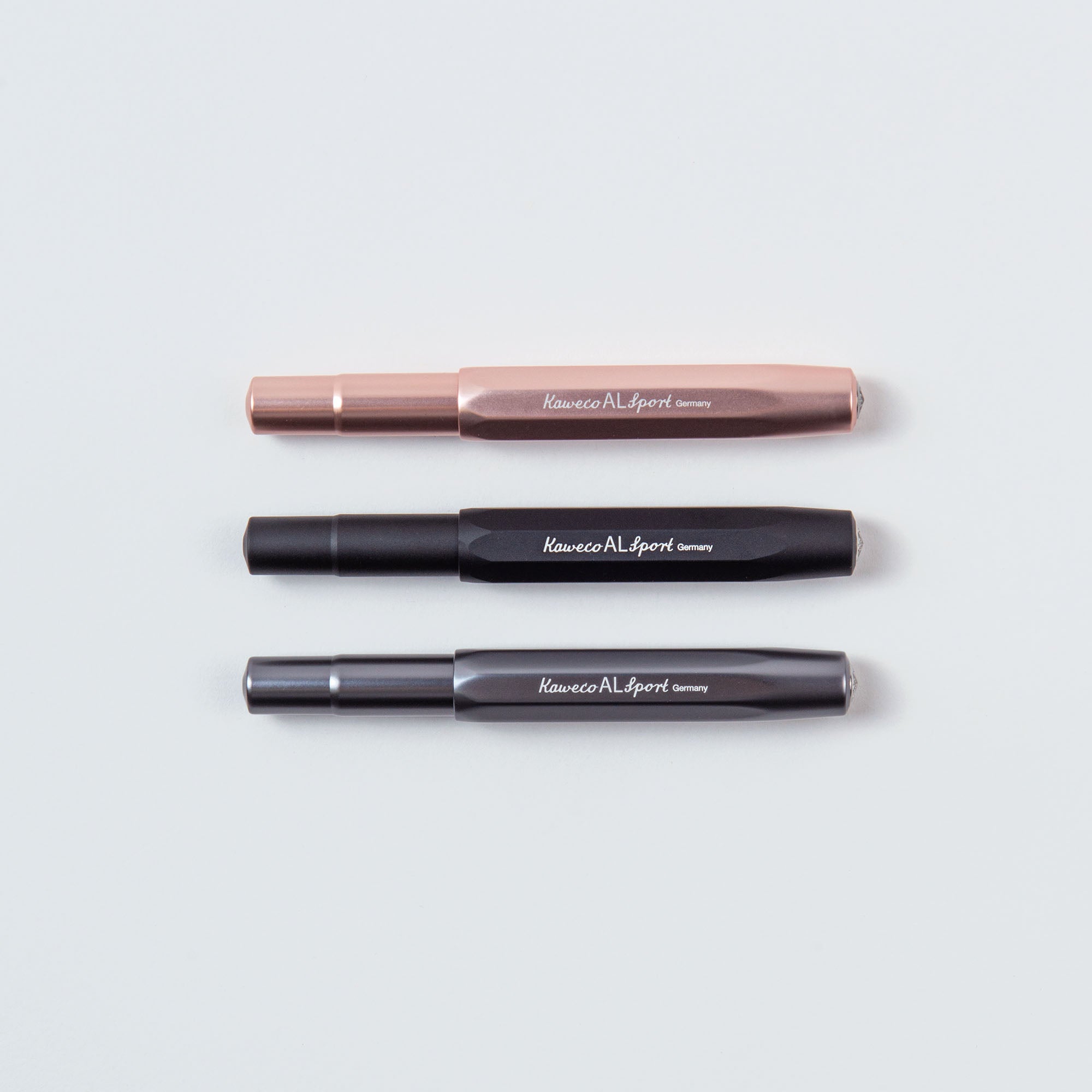 Drei Kaweco Füllhalter M – Aluminiumstifte, gelobt für ihre Kompaktgröße und ihr klassisches Design, liegen vor einem hellen Hintergrund. Sie sind horizontal ausgerichtet und zeigen elegante Oberflächen in Roségold, Schwarz und Grau.