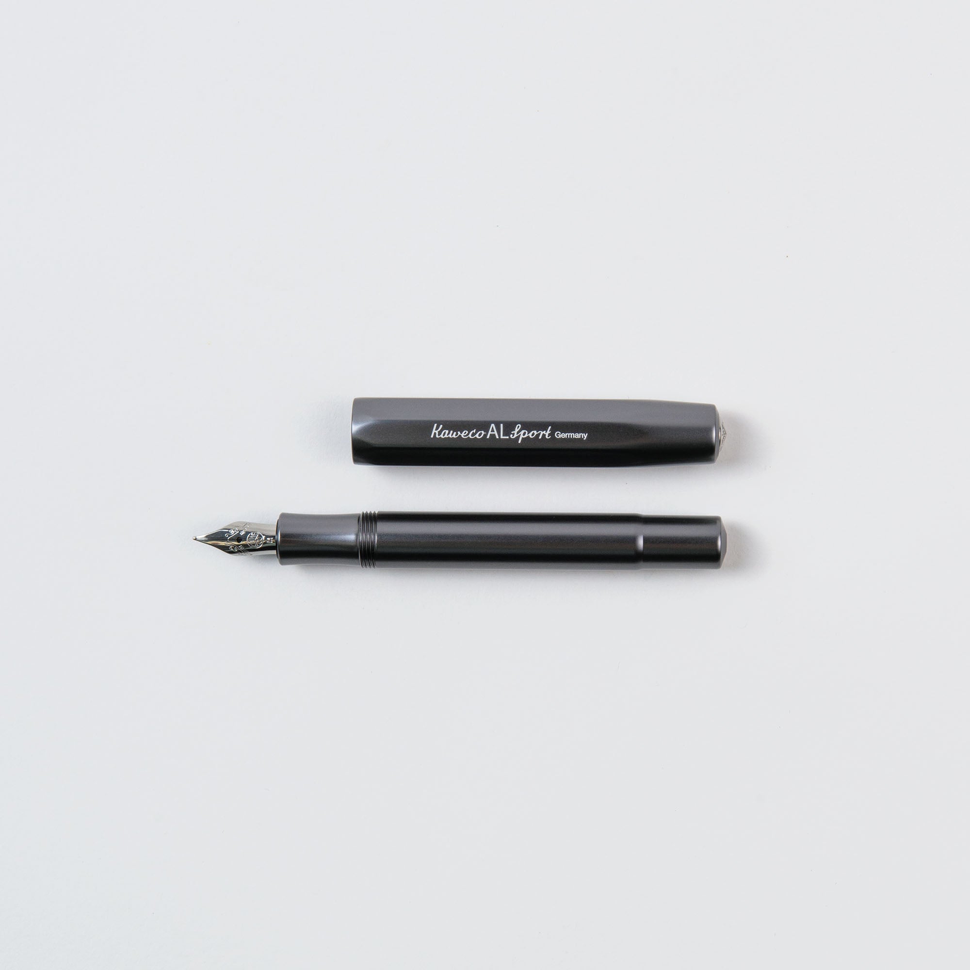 Ein schwarzer Kaweco Füllhalter M – Aluminium Füllhalter mit abnehmbarer Kappe liegt horizontal auf hellgrauem Untergrund und präsentiert seine Kompaktgröße. Die Kappe mit weißem Text liegt über dem Füllhalter und gibt den Blick auf die Feder frei.