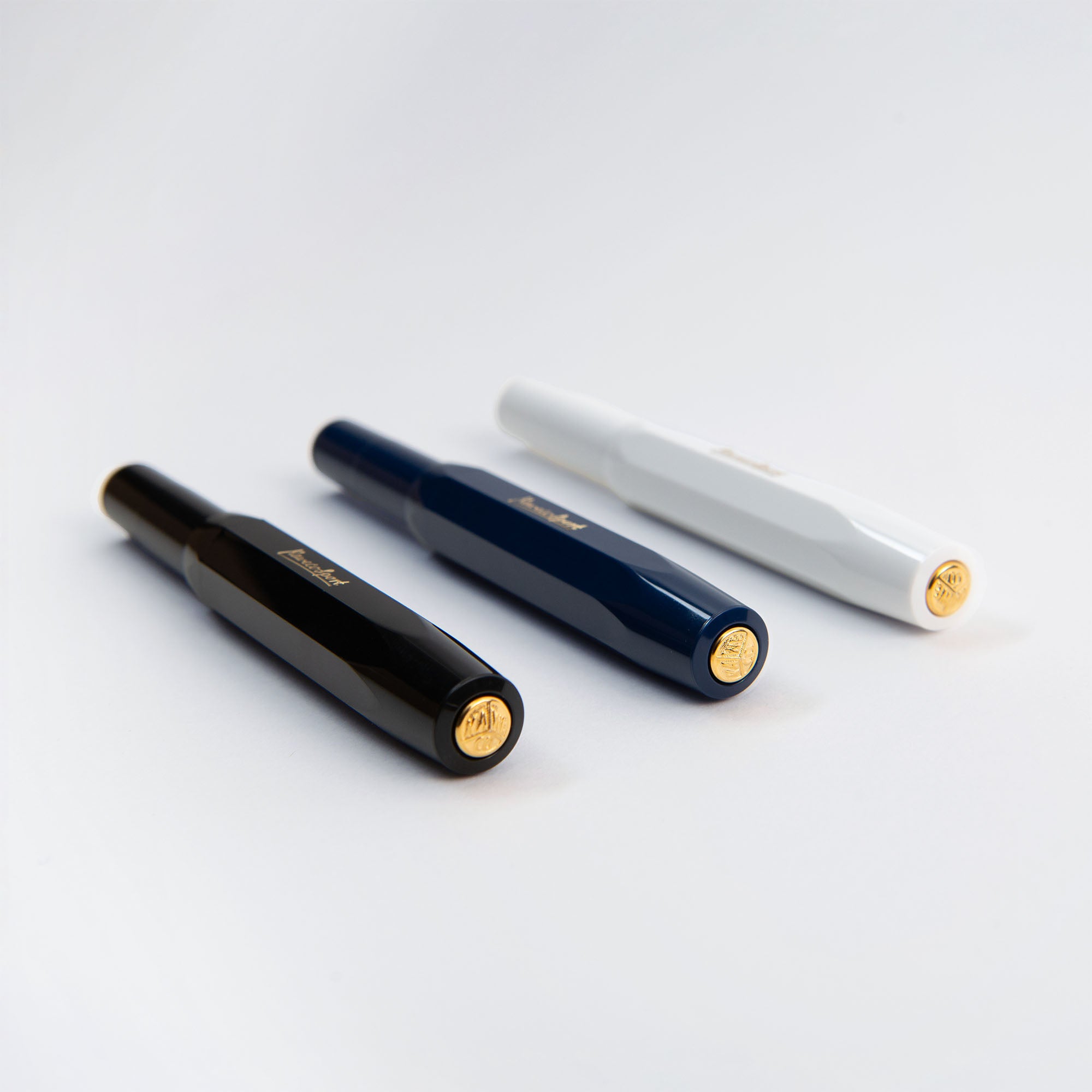 Drei Kaweco Classic Sport - Füllhalter von Kaweco in Schwarz, Marineblau und Weiß sind diagonal auf einer weißen Fläche angeordnet. Jeder Füllhalter hat eine Goldfeder und eine goldfarbene Kappe mit einem eingravierten Emblem.