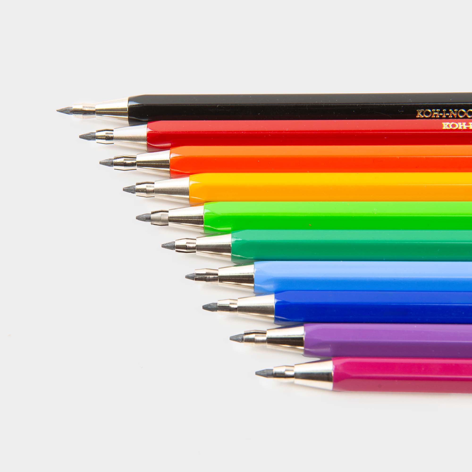 Eine Reihe farbiger Druckbleistifte Versatil von Koh-i-noor ist diagonal angeordnet, Spitzen links. Die 10 Farben - Schwarz, Rot, Orange, Gelb, Grün, Hellblau, Blau, Lila und Pink - warten auf einem weißen Hintergrund auf Ihr nächstes Meisterwerk.