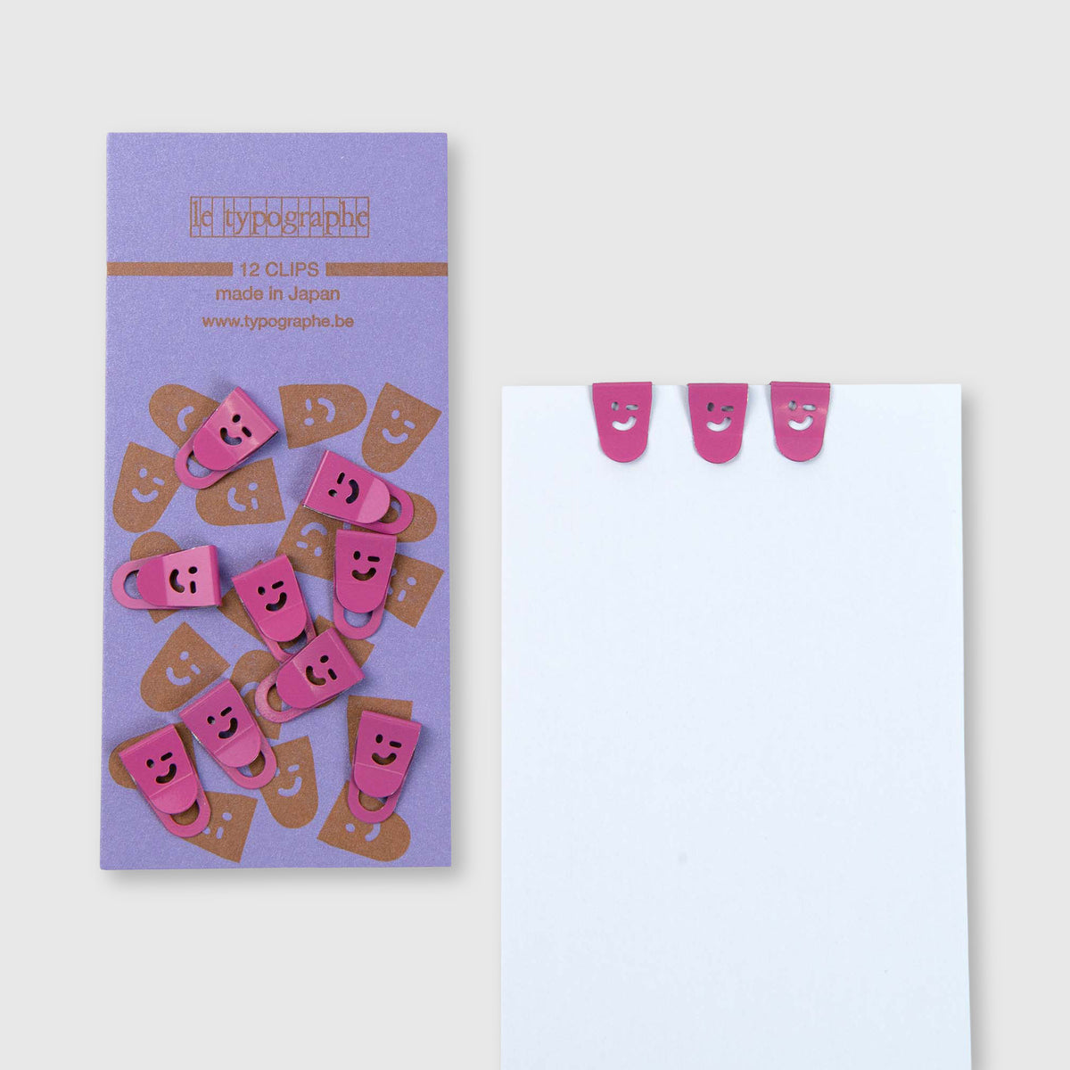 Auf einer violetten, rechteckigen Karte befinden sich zwölf rosa Metallklammern der Marke Le Typographe Mini Clips Smiley mit lächelnden Gesichtern, von denen drei oben an einem weißen Blatt Papier befestigt sind, um den Schreibtisch zu organisieren.