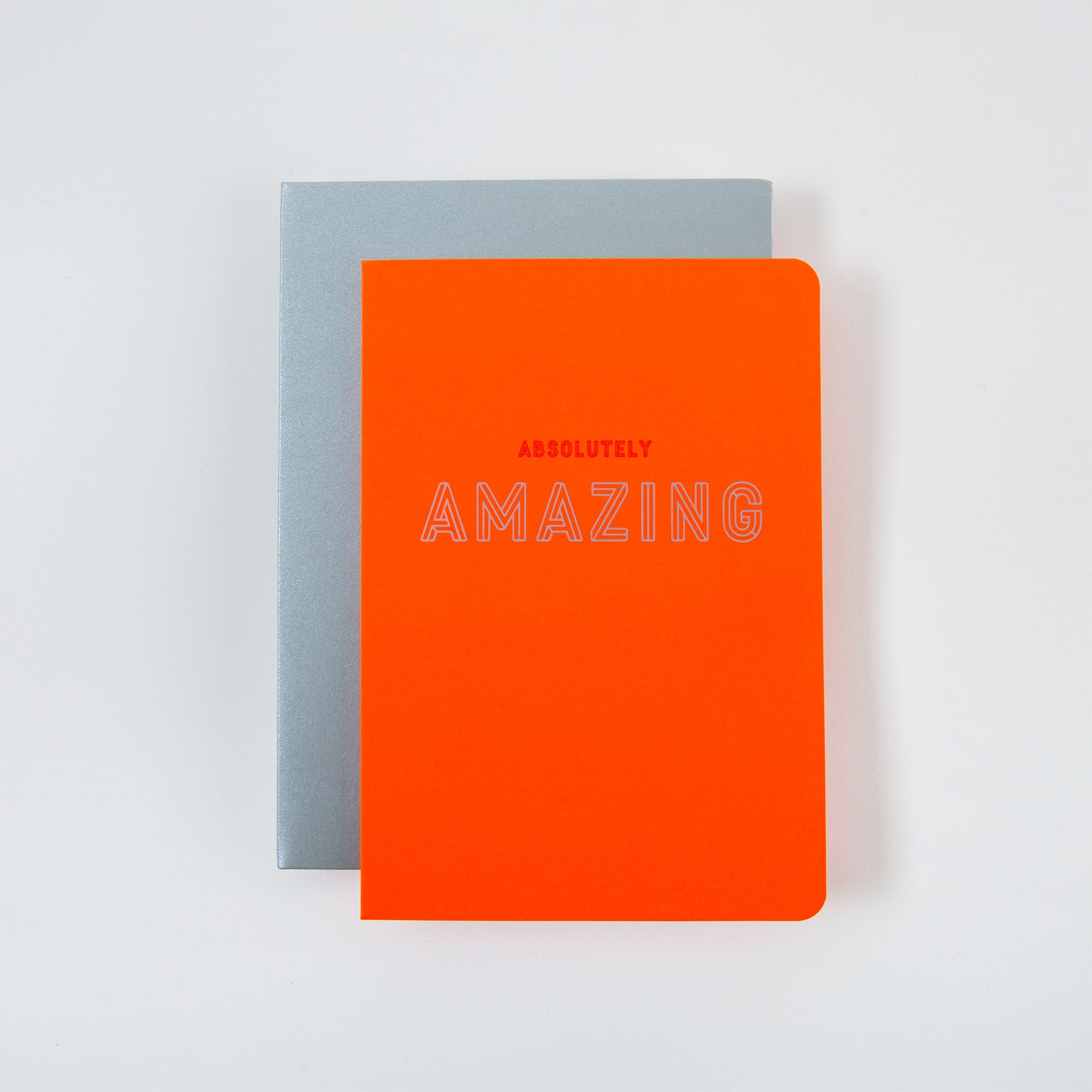 Auf einer weißen Oberfläche liegen zwei Notizbücher gestapelt. Das obere, von Le Typographe, ist leuchtend orange und trägt in reflektierender Schrift den Schriftzug „Absolutely Amazing“, der an Karte und Kuvert erinnert. Das untere Notizbuch ist schlicht hellgrau.