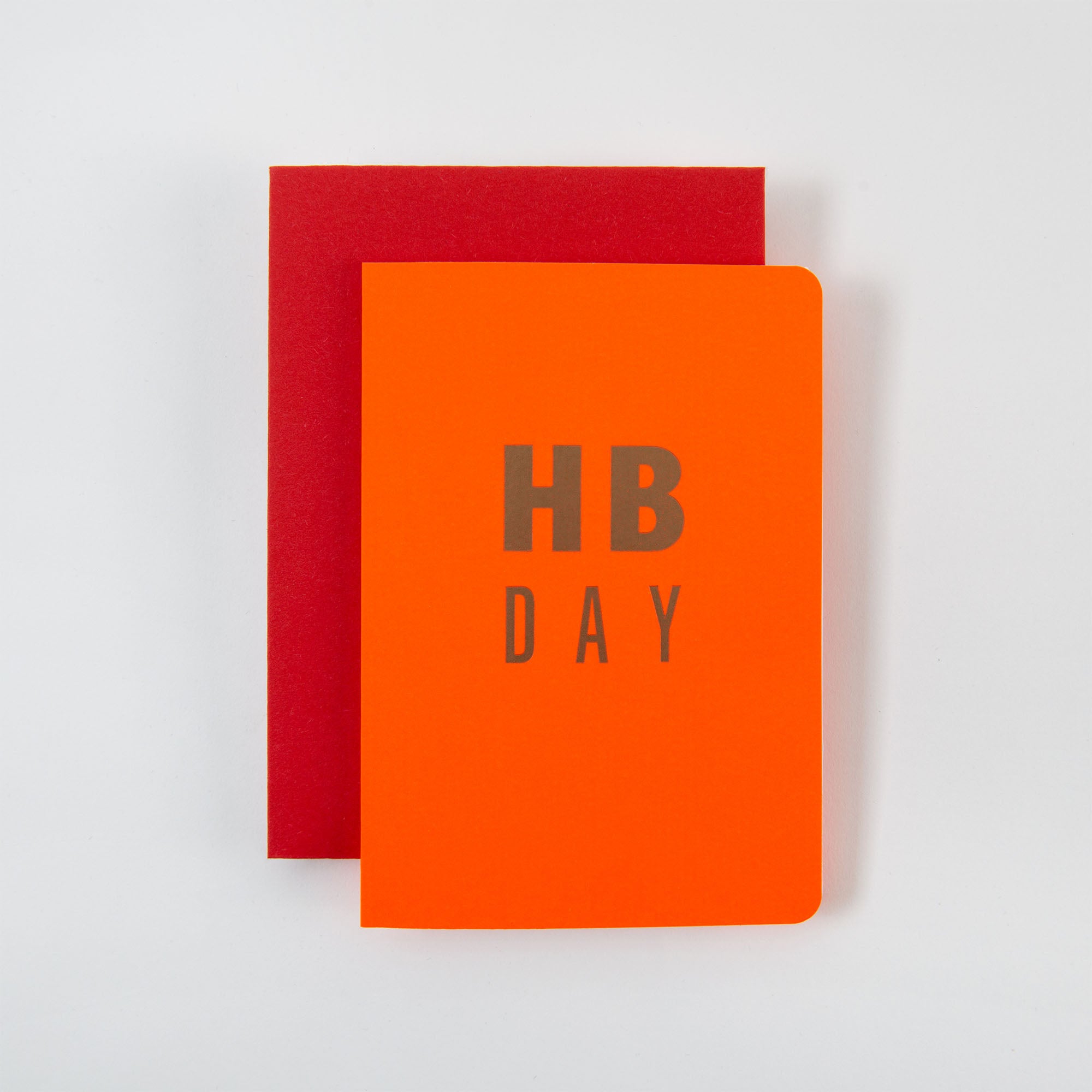 Ein roter Umschlag ist teilweise hinter einer orangefarbenen Le Typographe HB DAY Karte und einem Kuvert mit abgerundeten Ecken versteckt, auf deren Vorderseite „HB DAY“ in grauen Großbuchstaben steht. Diese Artikel sind ideal für eine Geburtstagsfeier und werden vor einem schlichten weißen Hintergrund präsentiert.
