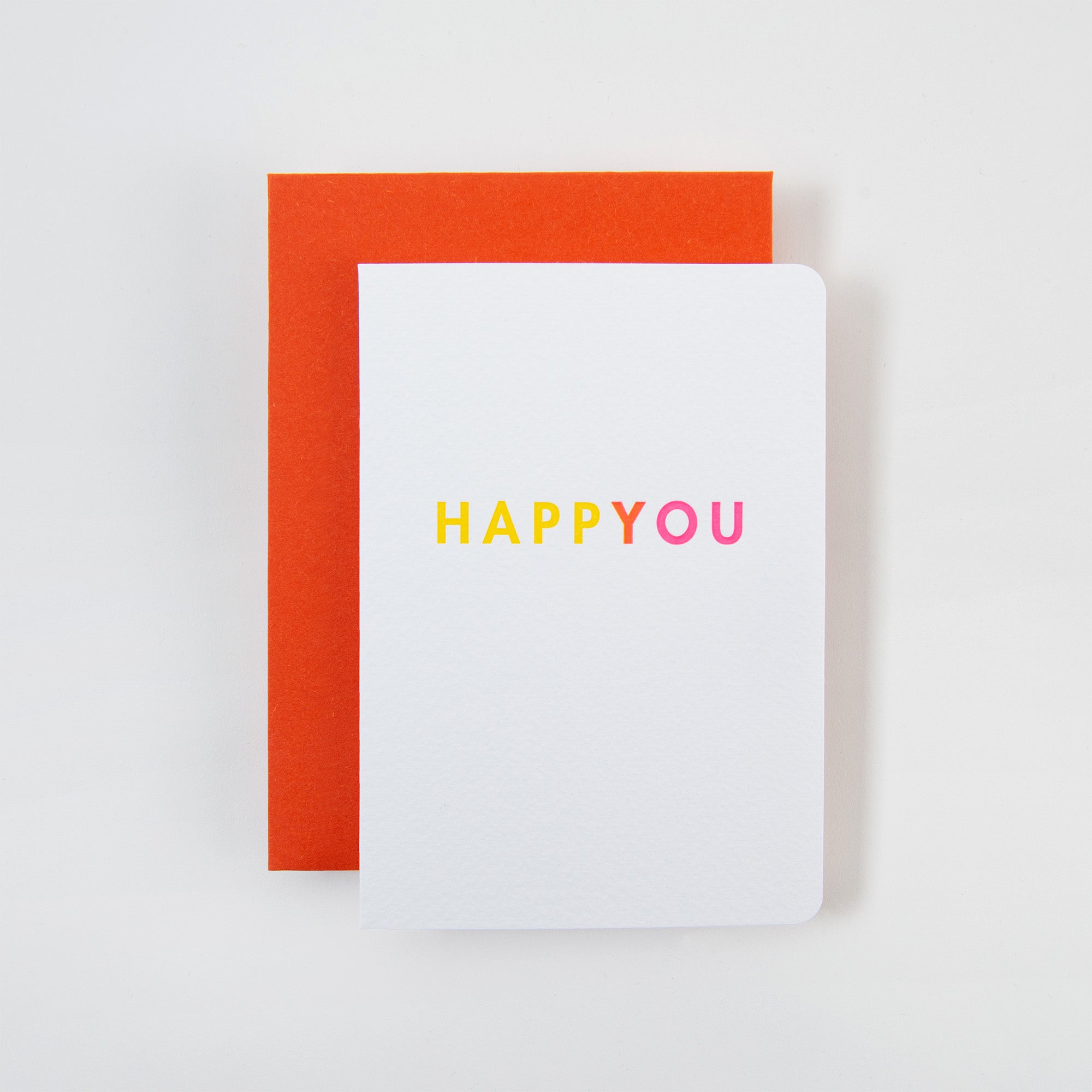 „HAPPYOU Karte und Kuvert“ von Le Typographe besteht aus einer weißen Klappkarte mit „HAPPY YOU DAY“ in Gelb und Pink, elegant platziert auf einem orangefarbenen Umschlag auf einer makellos weißen Oberfläche – ideal, um Ihren Lieblingsmenschen zu feiern.