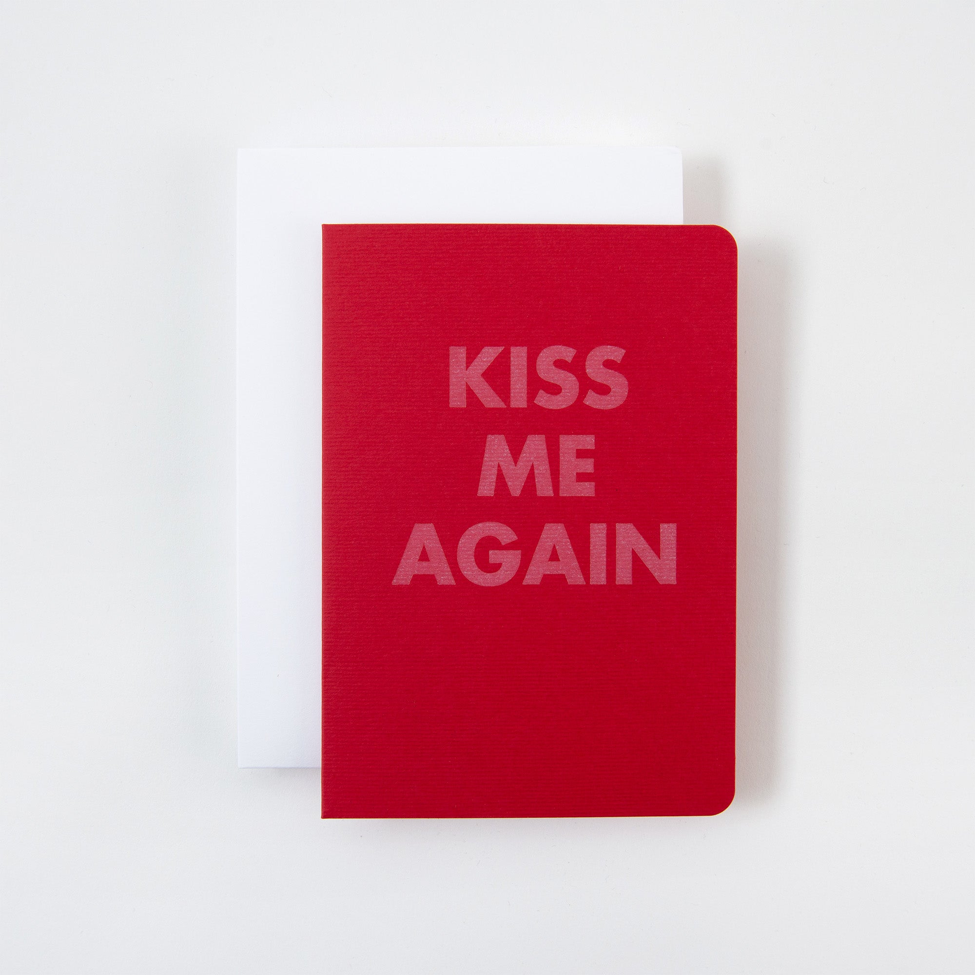 Eine bezaubernde rote Klappkarte von Le Typographe mit abgerundeten Ecken und dem fettgedruckten weißen Text „KISS ME AGAIN“ ruht elegant auf einer schlichten weißen Karte vor einem hellen Hintergrund, perfekt zum Valentinstag.