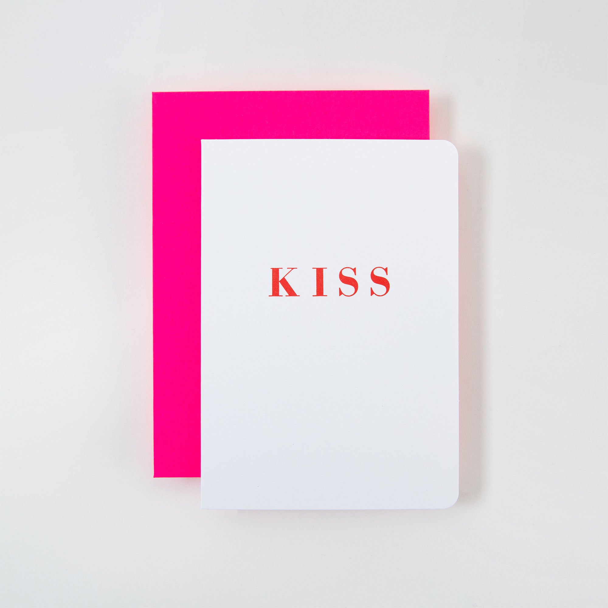 Ein weißes Notizbuch mit abgerundeten Ecken, auf dem in kräftigen roten Buchstaben „KISS“ steht, liegt auf einer großen, leuchtend rosa KISS Karte und Kuvert von Le Typographe. Der schlichte weiße Hintergrund betont die lebendige Kombination.