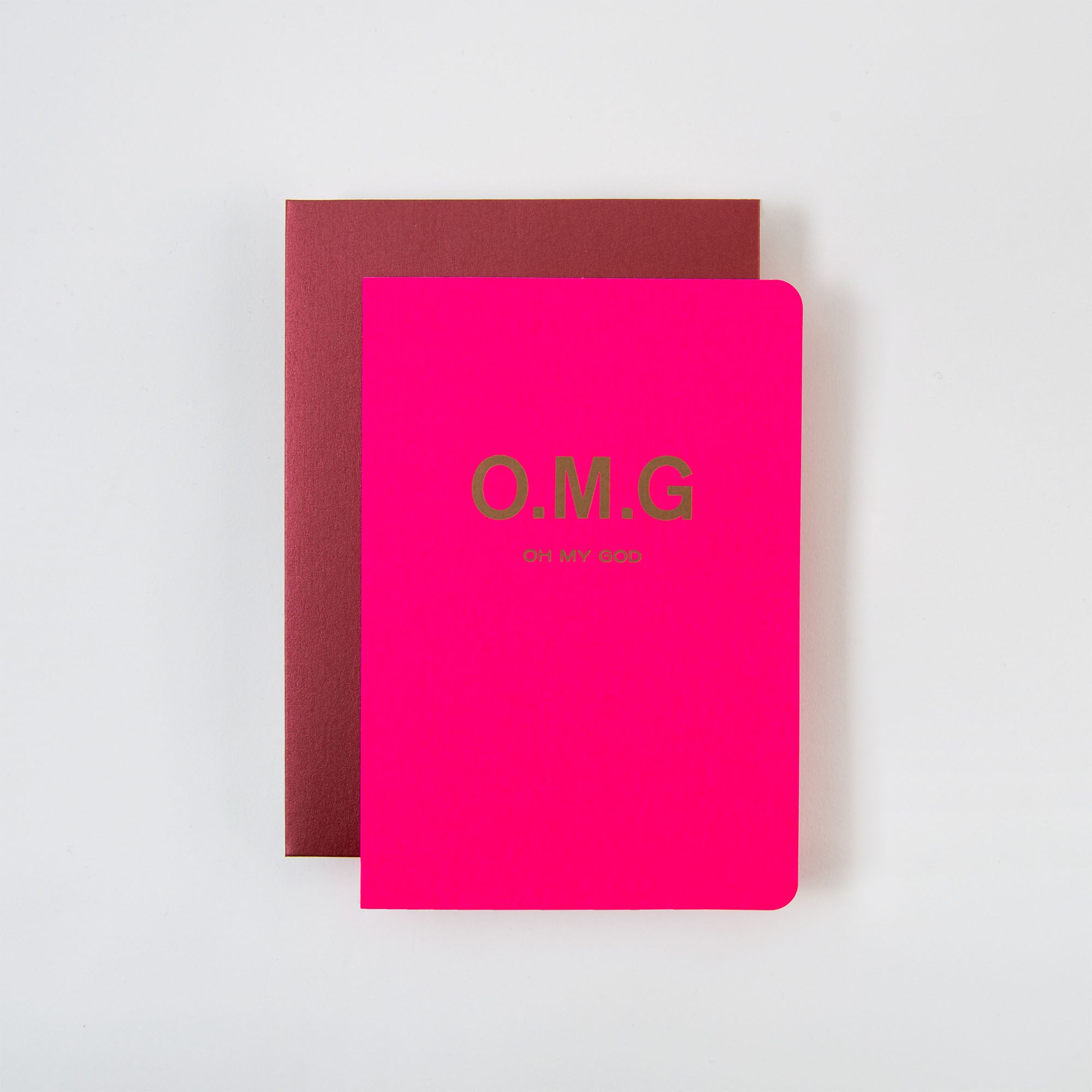 Eine leuchtend rosa „OMG Karte und Kuvert“ von Le Typographe mit den goldenen Aufschriften „O.M.G“ und „OH MY GOD“ liegt auf einem kastanienbraunen Notizbuch auf einer weißen Oberfläche und erzeugt so einen eindrucksvollen optischen Kontrast.