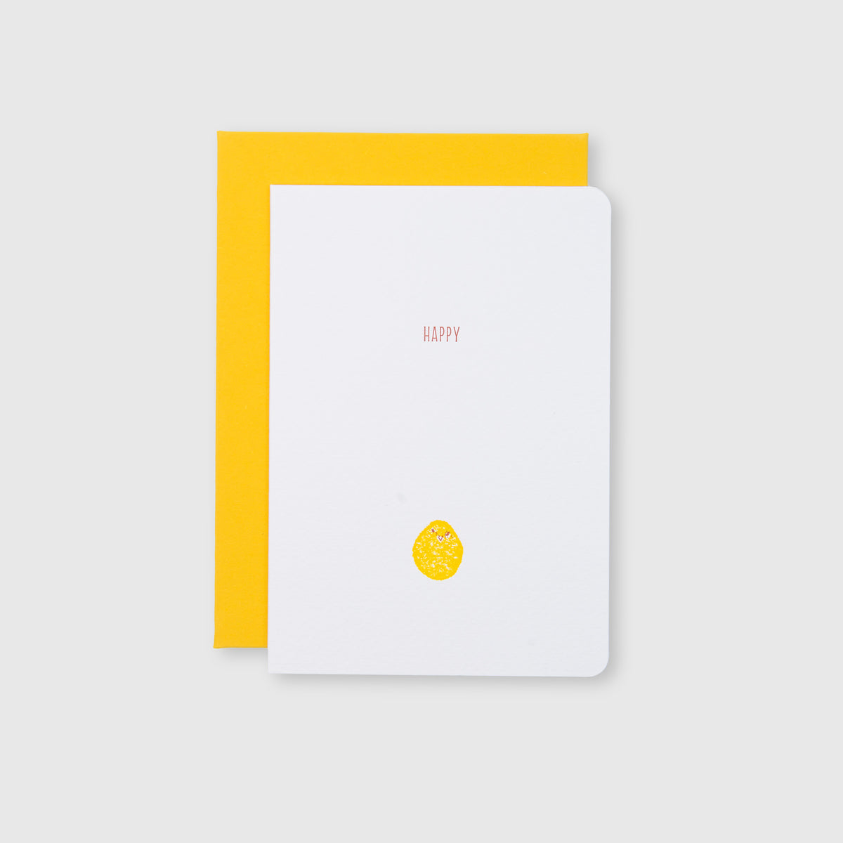 Eine weiße „CHICK“-Grußkarte mit Umschlag von Le Typographe, auf der in kleinen roten Buchstaben „HAPPY“ über einer gelben Zitronenillustration zu lesen ist, die im traditionellen Buchdruckverfahren hergestellt wurde, liegt auf einem leuchtend gelben Umschlag. Die Szene ist vor einem hellgrauen Hintergrund dargestellt.