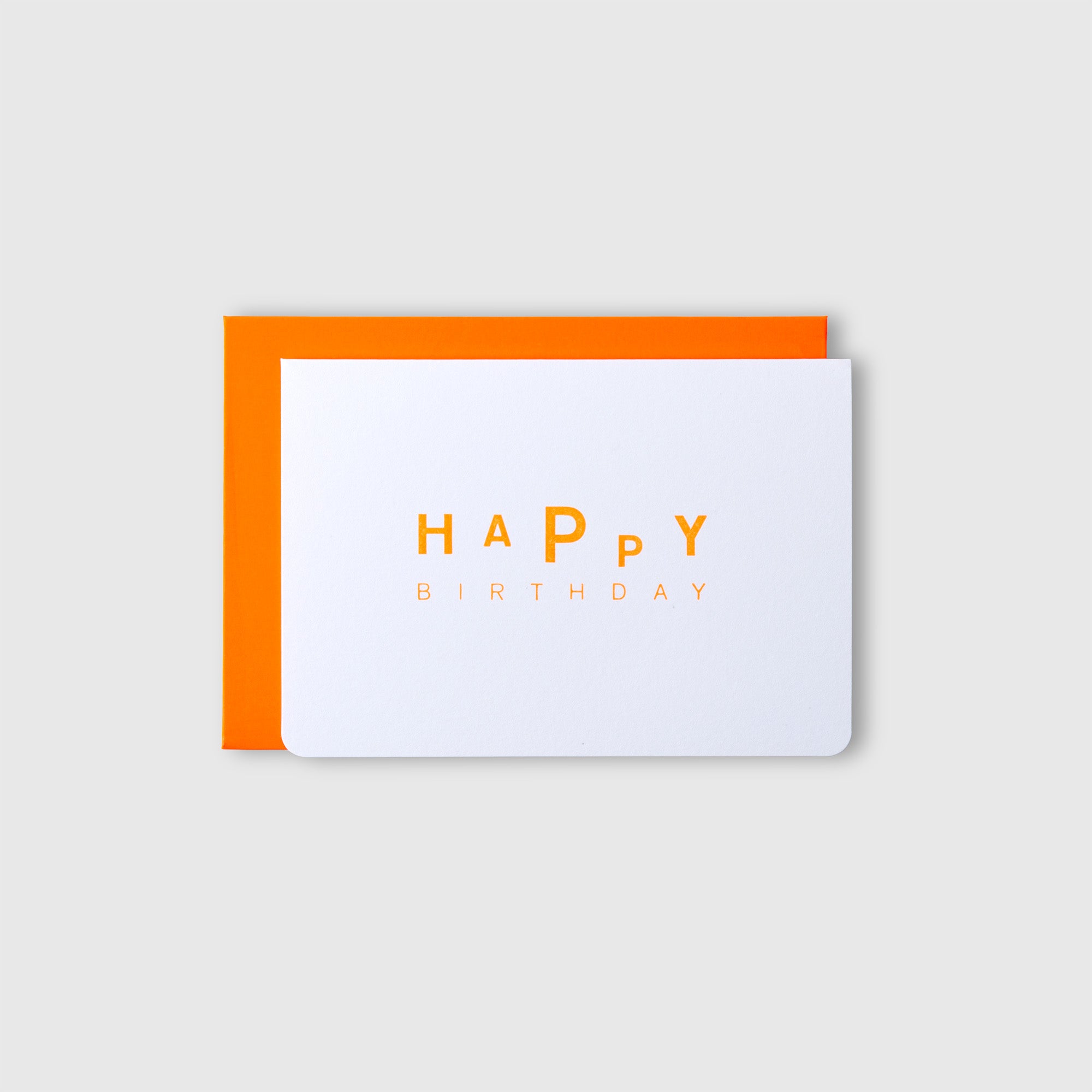 Eine weiße Letterpress-Karte „Dancing Birthday – Karte Kuvert“ von Le Typographe mit der Aufschrift „HAPPY BIRTHDAY“ in orangefarbenen und gelben Buchstaben liegt auf einem neonorangefarbenen Umschlag vor einem hellgrauen Hintergrund.
