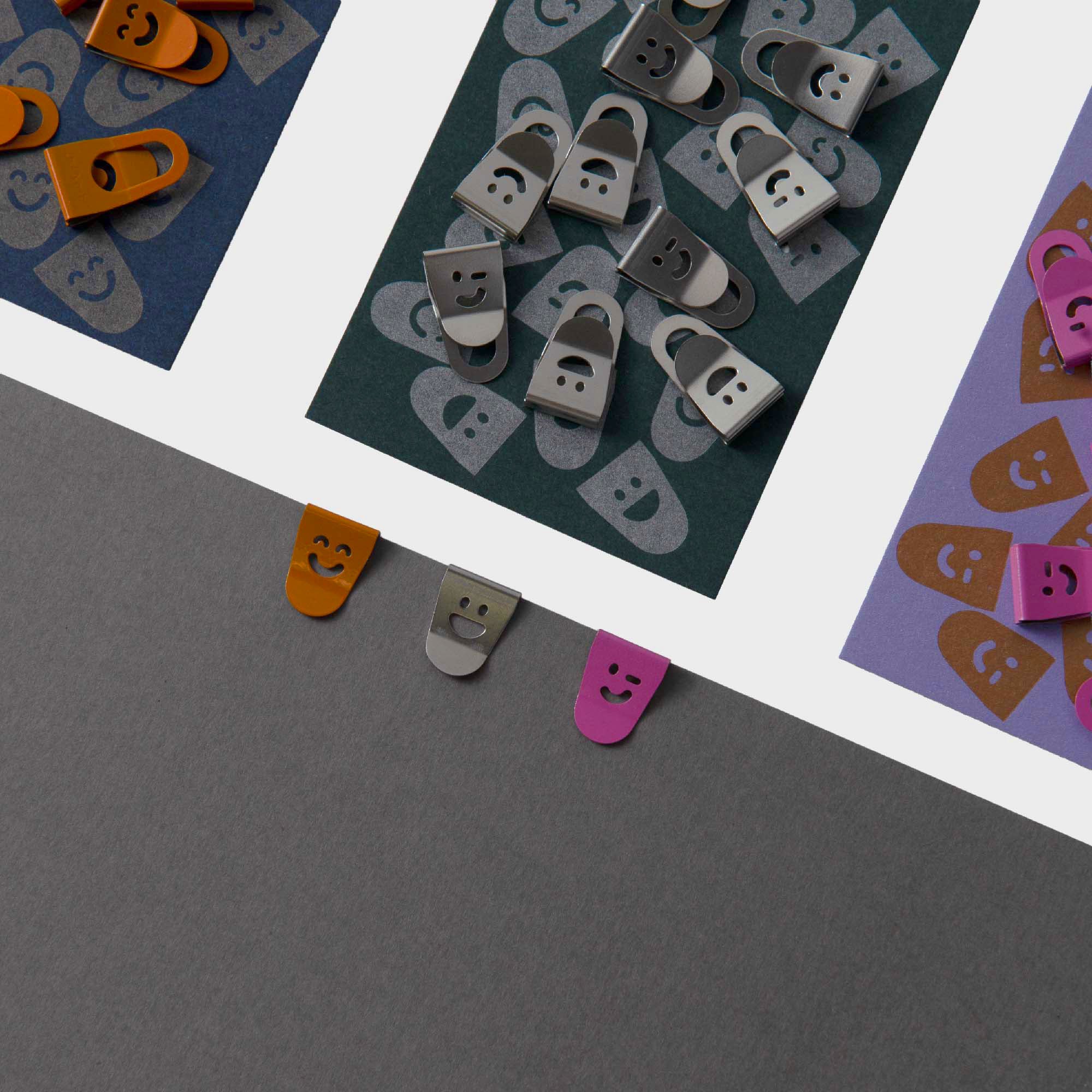 Die Mini Clips Smiley von Le Typographe sind nach Farbe und Smiley-Gesicht auf dunklem Papier sortiert, wobei einige auf ein graues Blatt geklippt sind - perfekt, um Ihren Schreibtisch zu organisieren.