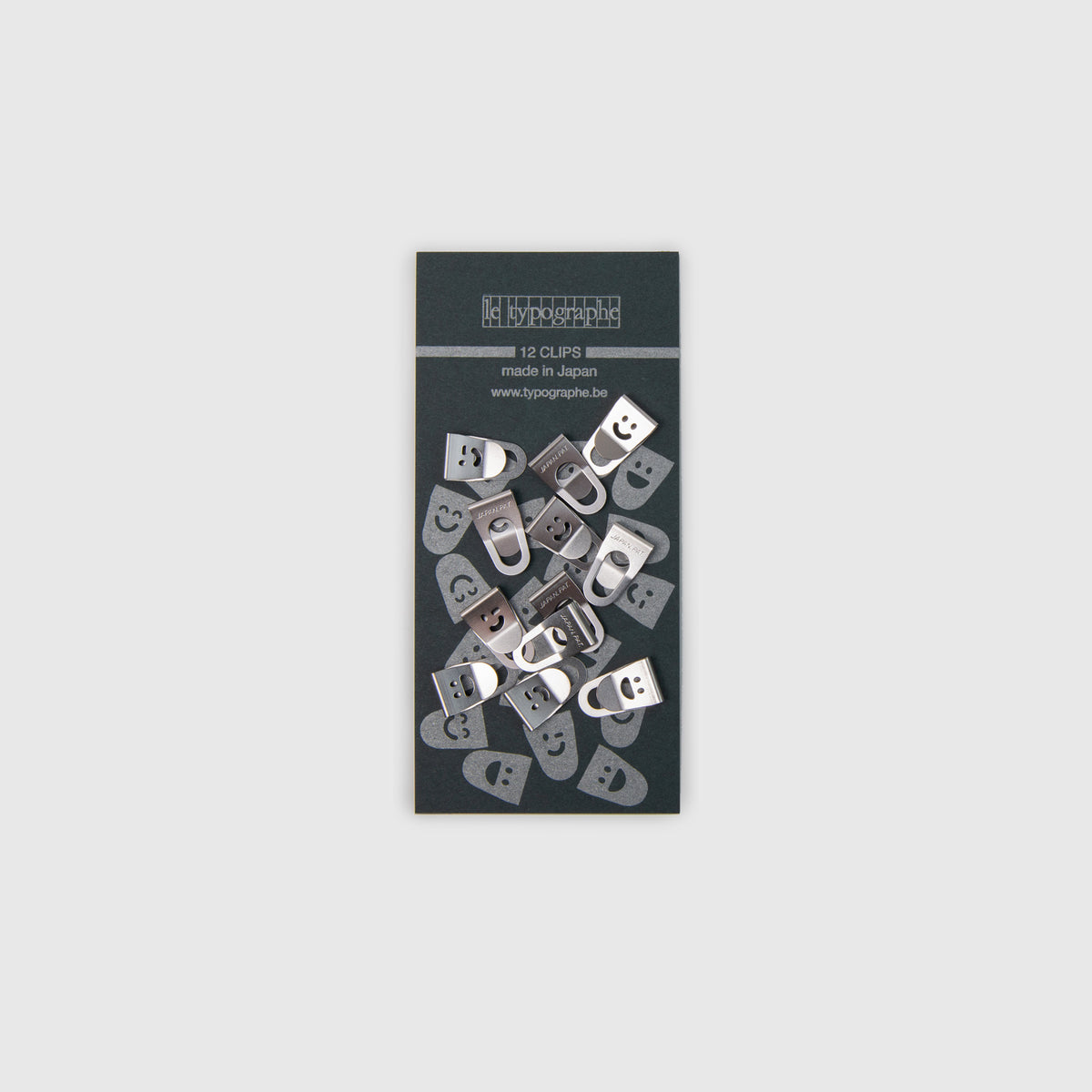 Eine Packung mit zwölf silbernen Metallklammern Le Typographe Mini Clips Smiley, die jeweils mit einem Smiley-Gesicht geprägt sind und auf einer dunkelgrauen Trägerkarte befestigt sind.