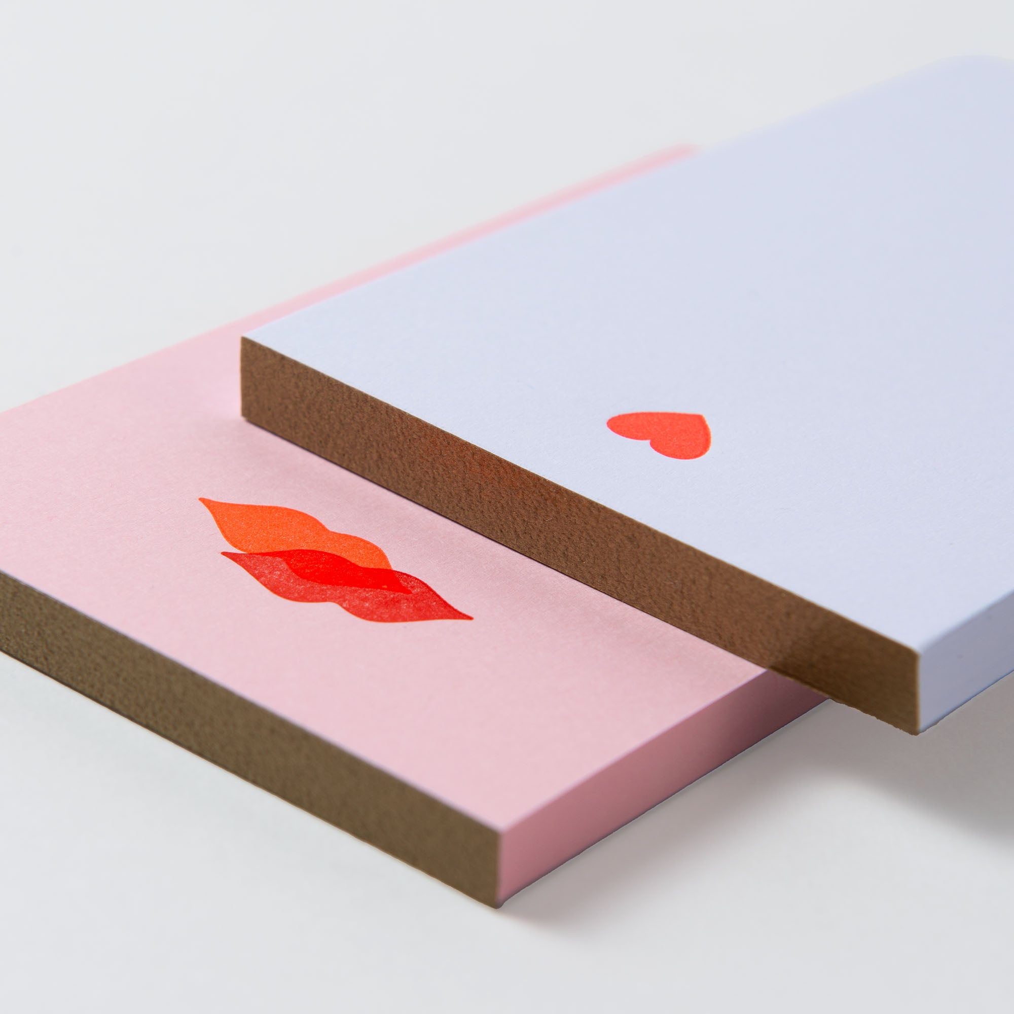 Zwei Mini-Notizblöcke - Kiss Kiss, Love Love von Le Typographe kreuzen sich auf einer Oberfläche. Der rosafarbene hat einen roten Lippenaufdruck und der hellblaue hat oben ein kleines rotes Herz. Ihr braun umrandeter PapierLetterpress-Stil ist einfach bezaubernd.