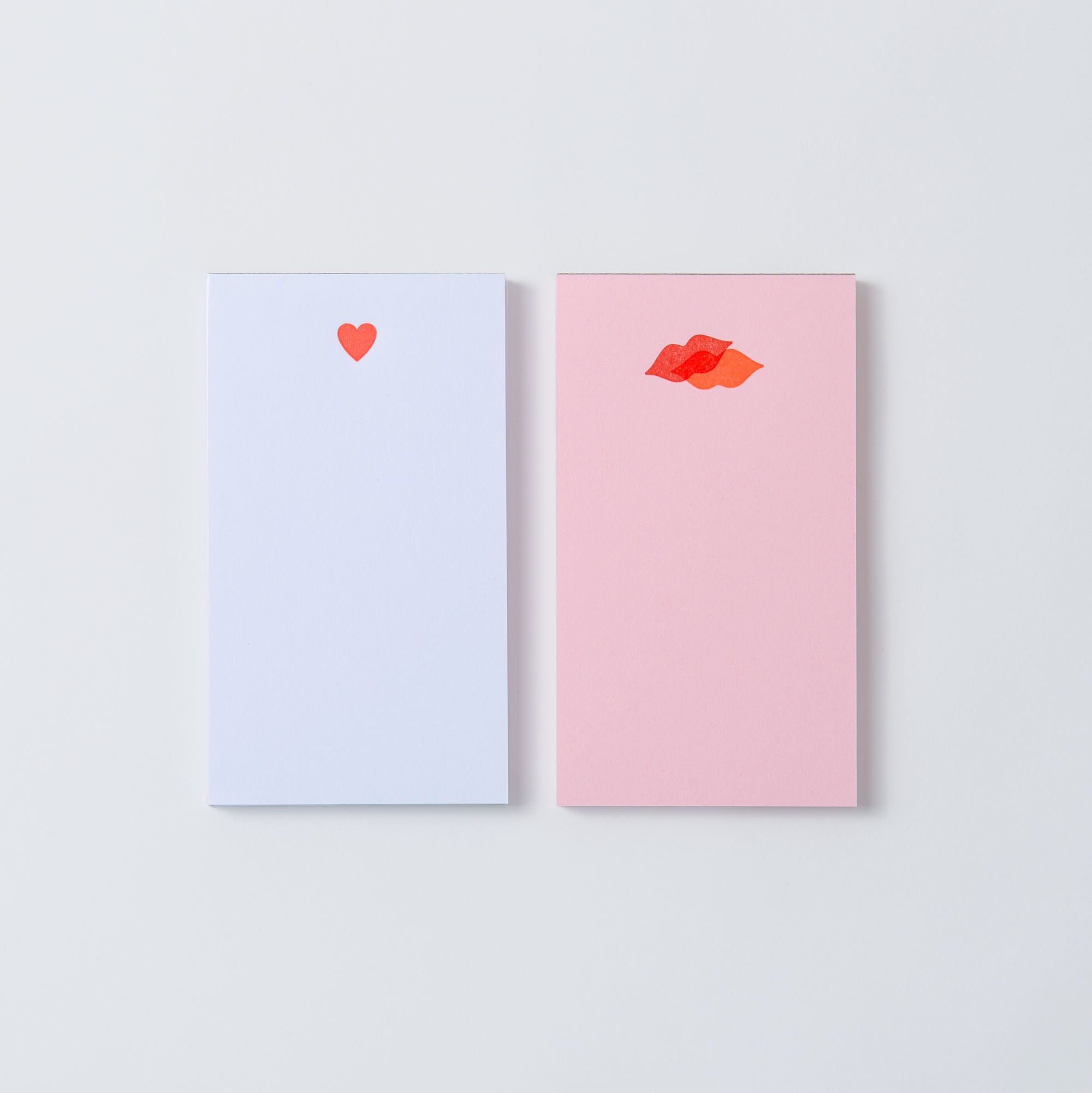 Zwei Mini-Notizblöcke aus der Kollektion „Mini Notepads – Kiss Kiss, Love Love“ von Le Typographe liegen auf einem weißen Hintergrund. Einer ist weiß mit einem kleinen roten Herz, während der andere rosa mit winzigen roten Lippen ist. Beide wurden mit PapierLetterpress hergestellt.