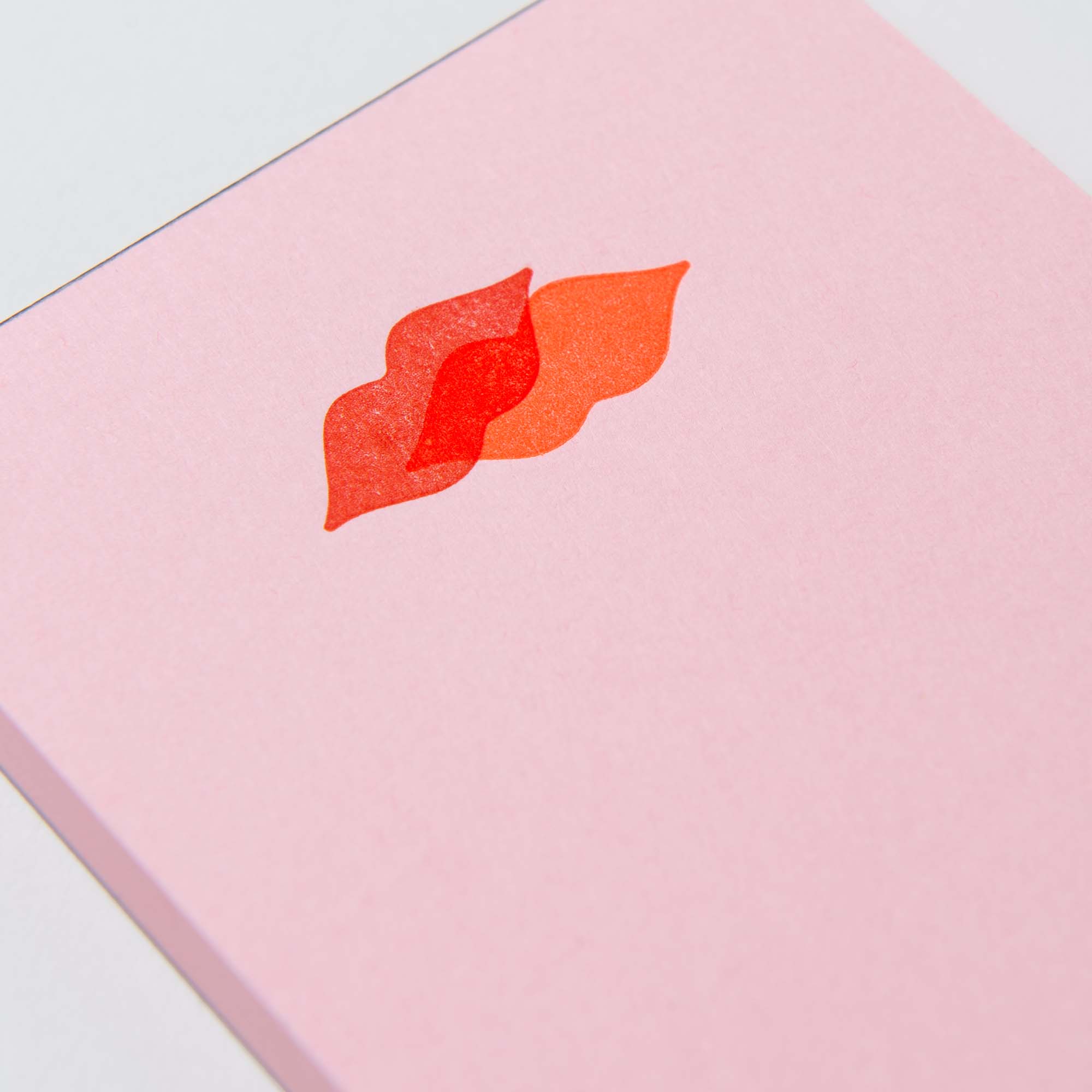 Der Mini-Notizblock „Kiss Kiss, Love Love“ von Le Typographe hat einen rosa Einband mit einem einzigartigen stilisierten Design aus überlappenden roten und orangefarbenen Lippen am oberen Rand und ist auf den Abreißseiten mit Papierletterpress-Details versehen.